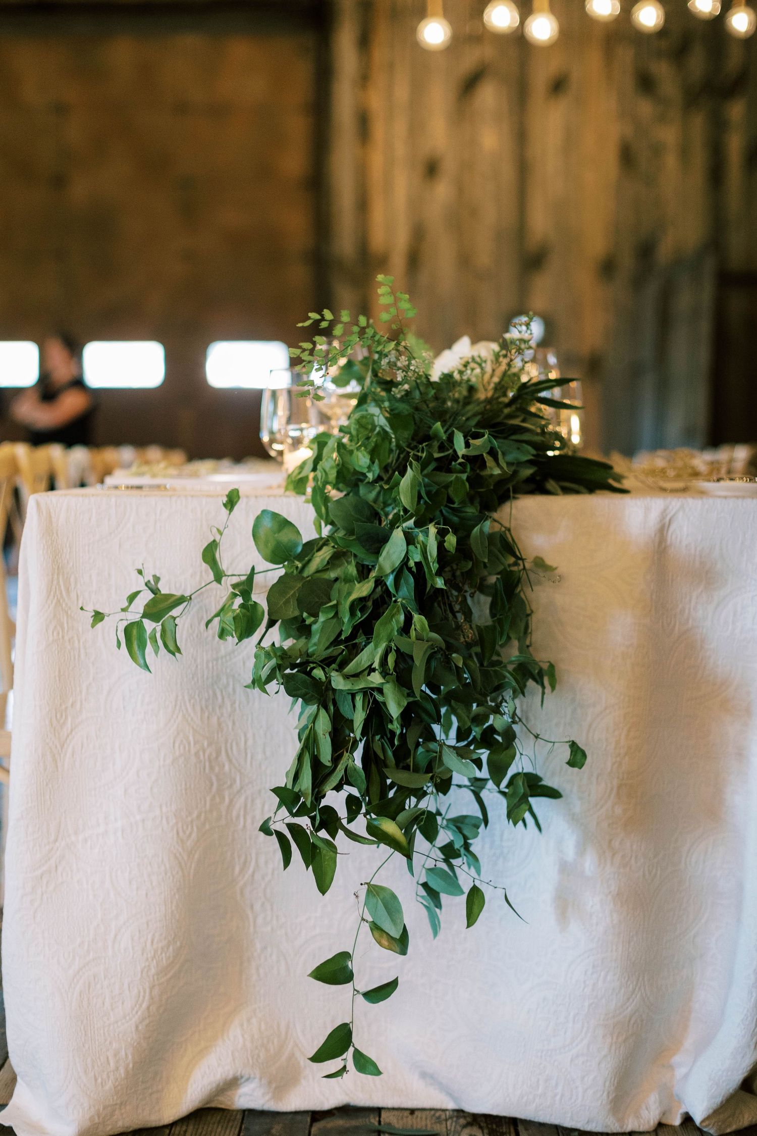Lush green foliage arrangement cascading over white tablecloth at wedding reception.