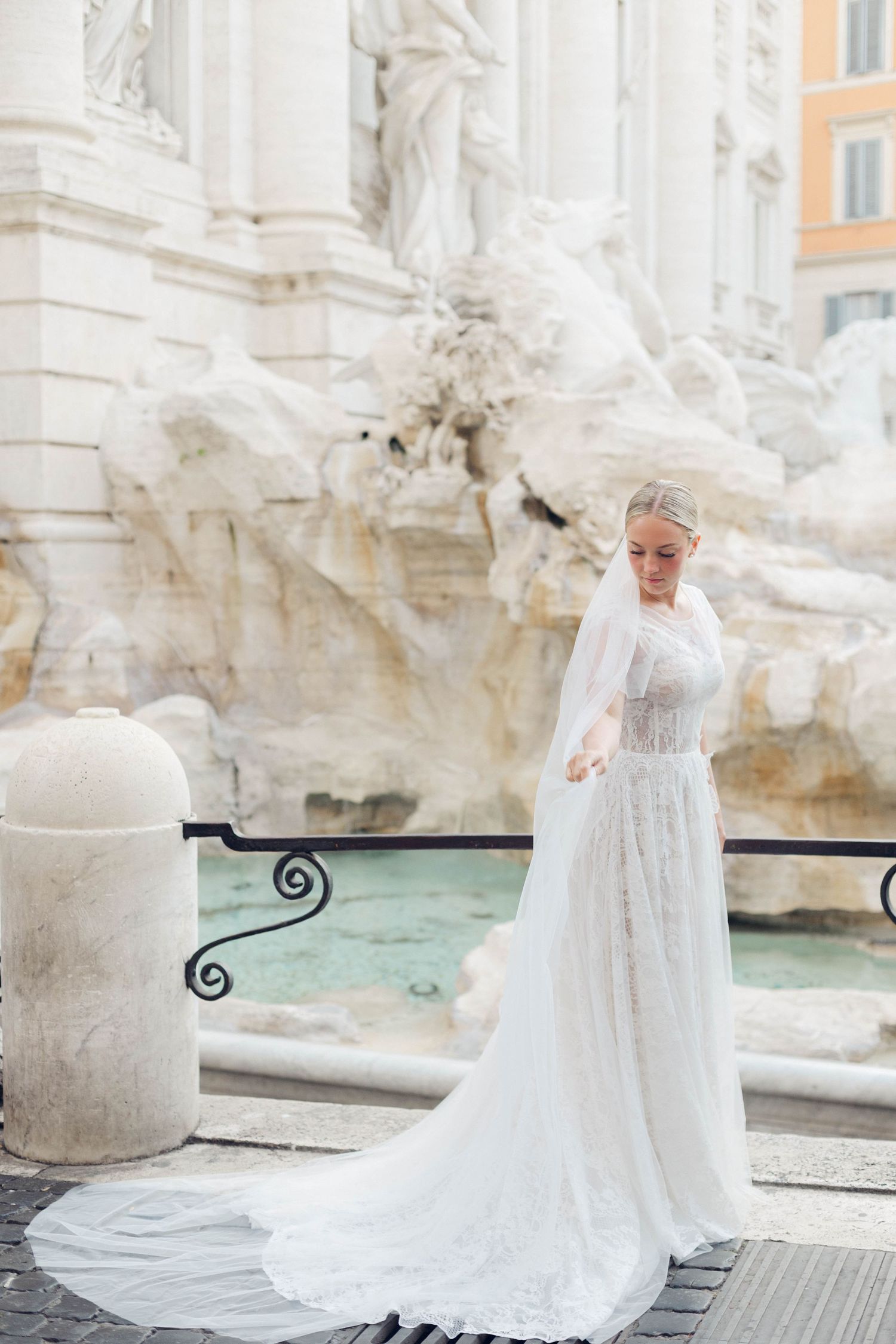 Bride in flowing lace dress poses by the Trevi Fountain with cascading waters in background.