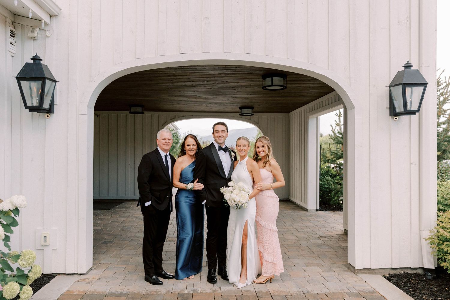 Group photo of wedding party standing under white archway with lanterns.
