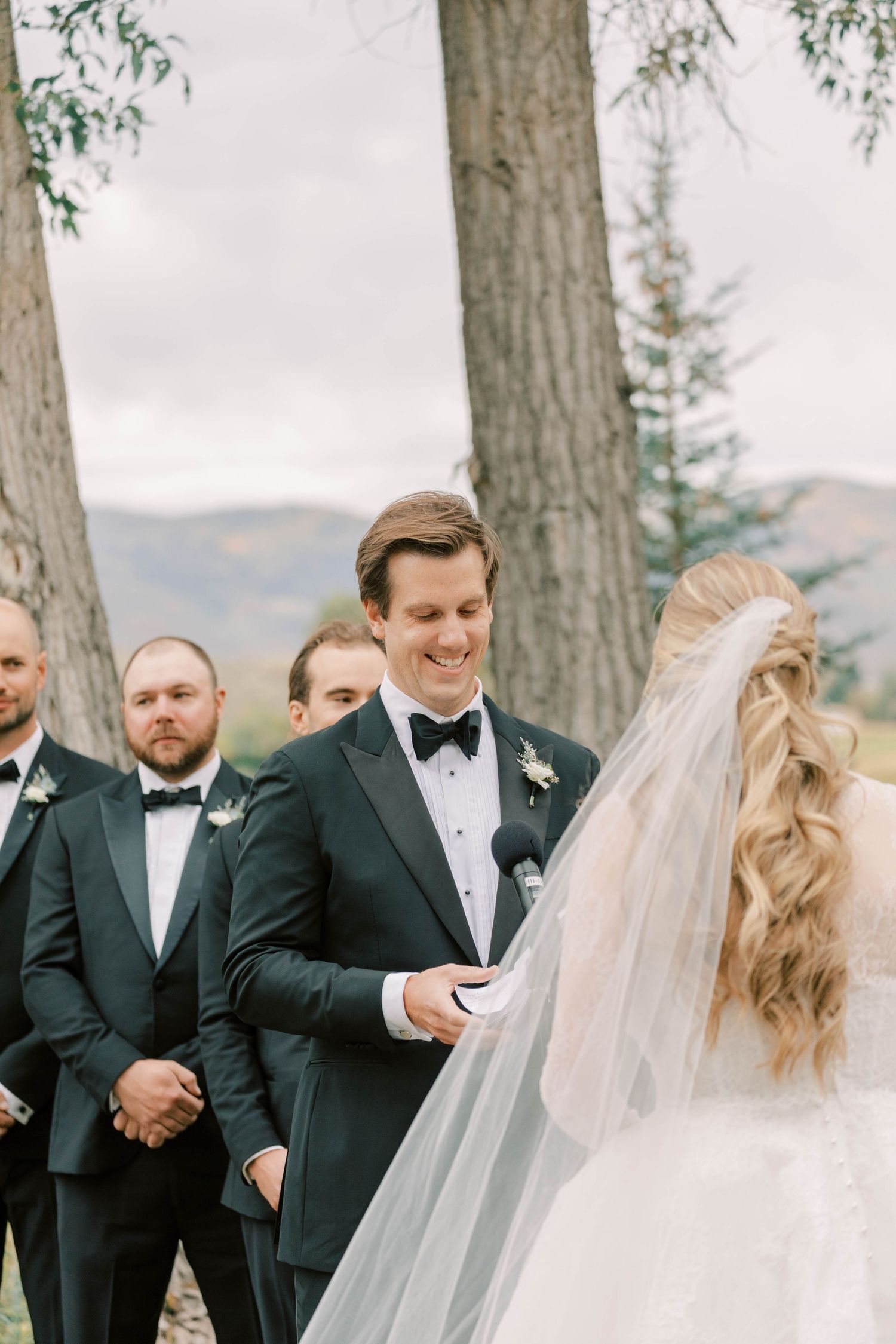 A couple exchanges vows during outdoor mountain wedding ceremony with groomsmen standing nearby.