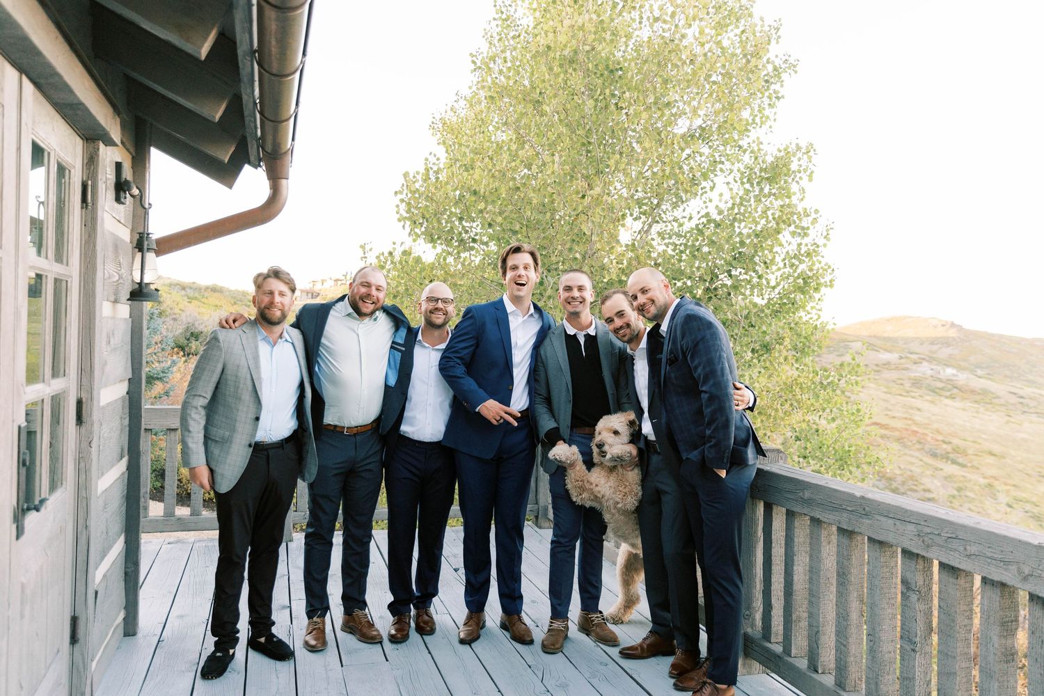 Group of formally dressed people and a goldendoodle dog gather on an outdoor deck.