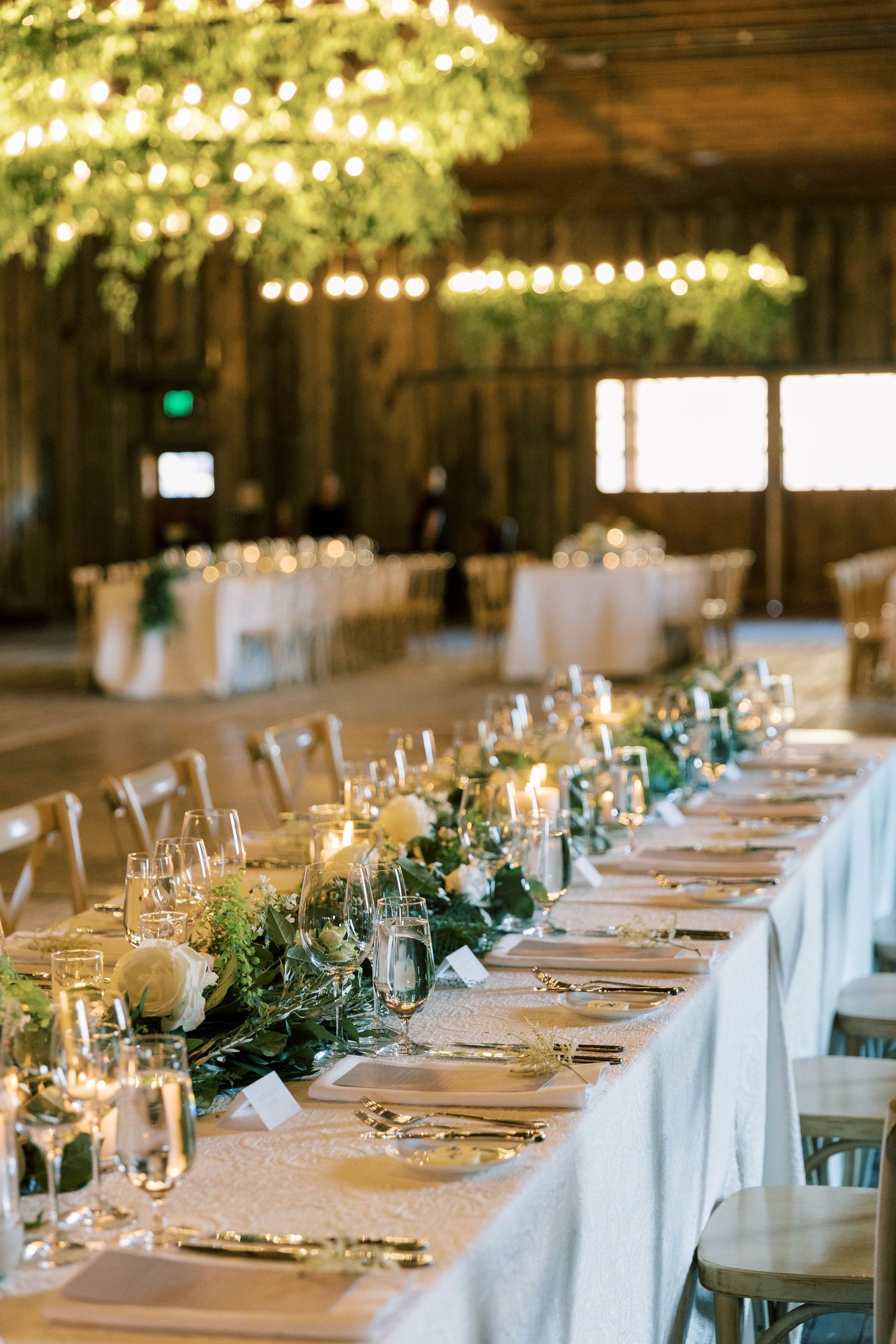 Elegant wedding reception table setup with white linens, greenery centerpieces and string lights in a rustic barn venue.