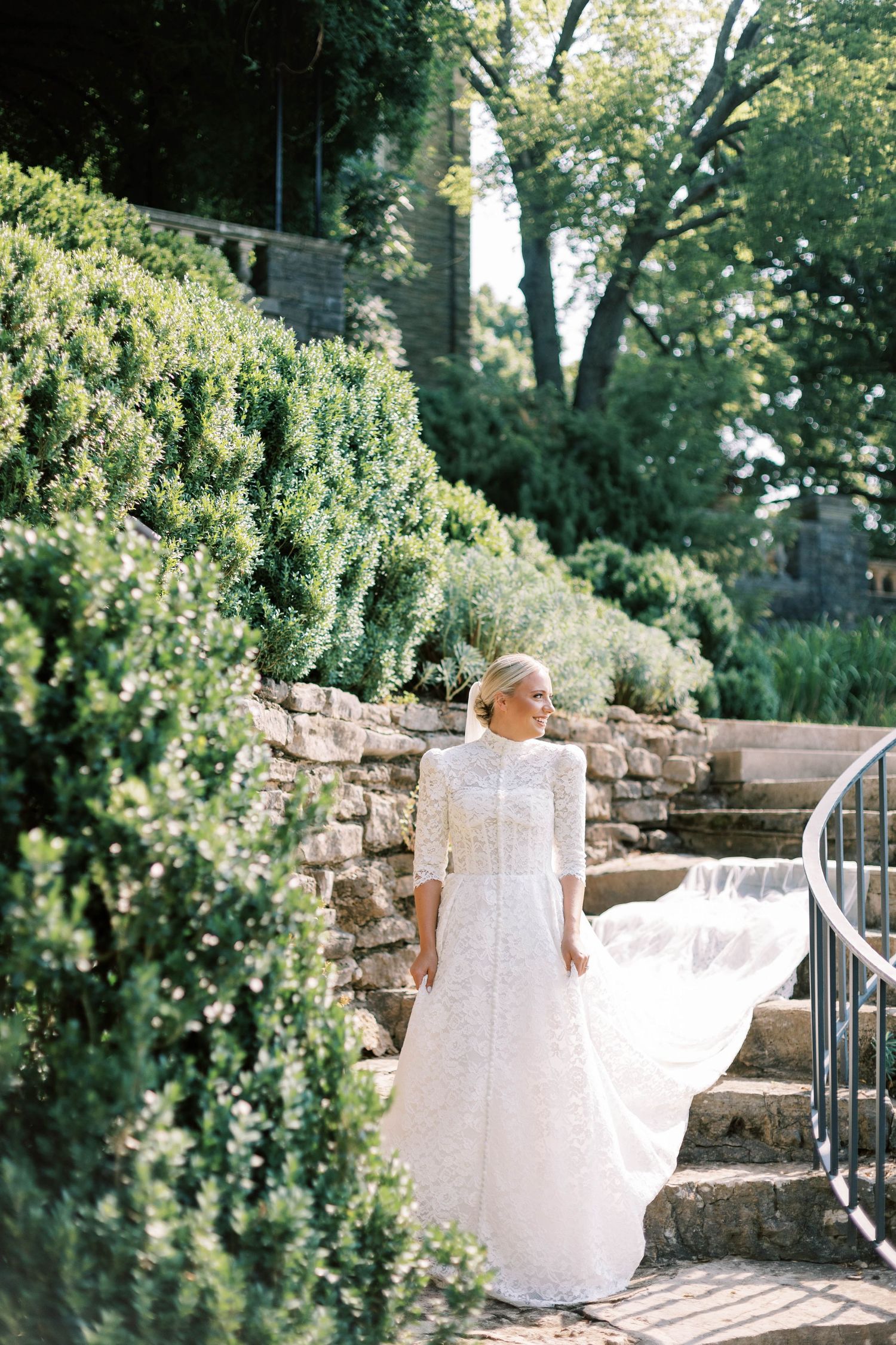 White wedding dress flows gracefully on stone garden steps amid green shrubbery at Cheekwood Estate.