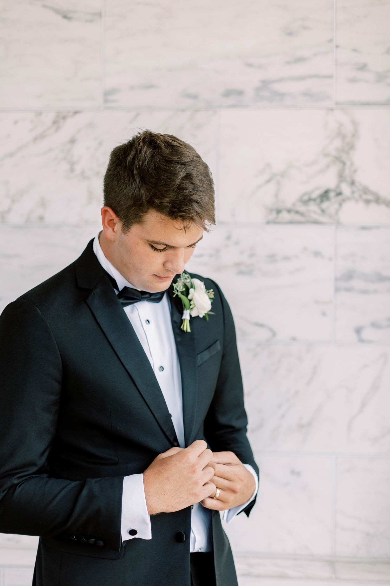 Groom in classic black tuxedo adjusts his cuff links against marble wall backdrop with white boutonniere.