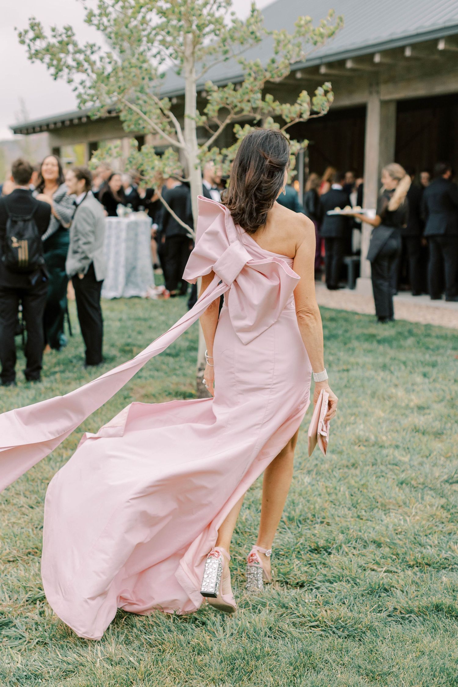 Woman in flowing pink gown twirling on grass during outdoor wedding celebration.