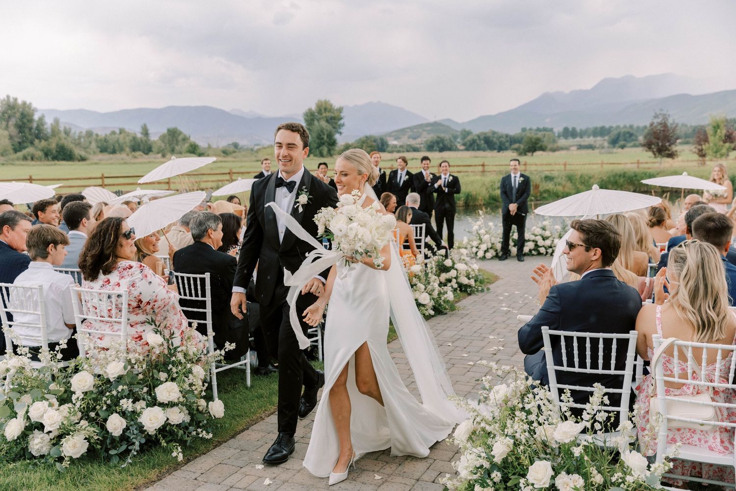 Happy couple walking down aisle after ceremony between white chairs and floral arrangements.