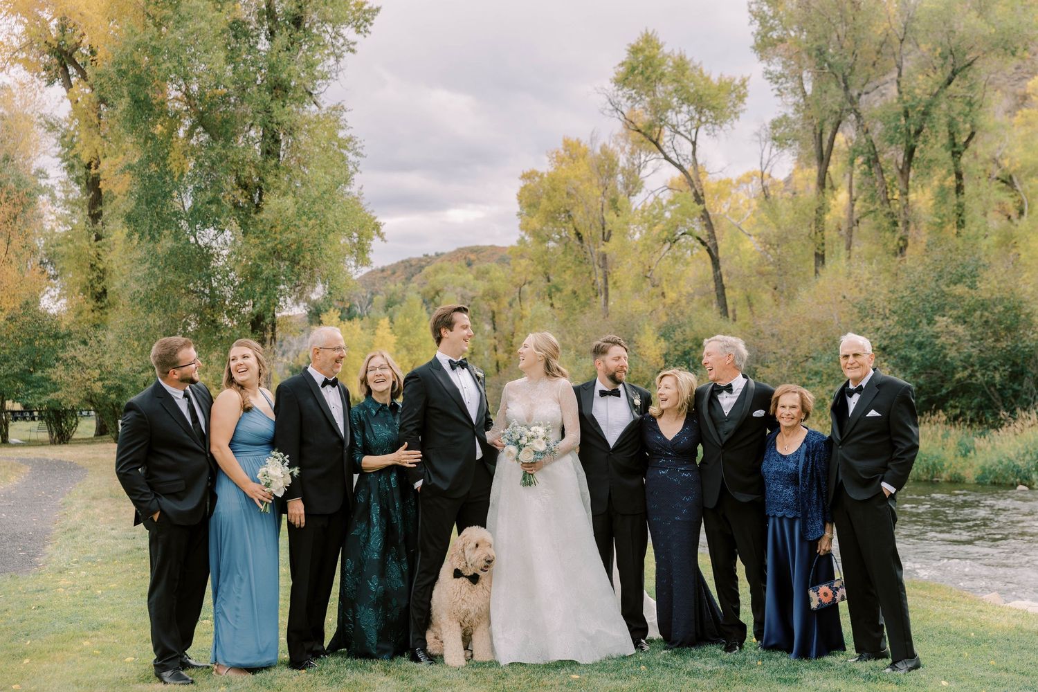 Wedding party and family members gather for formal group photo with dog by river.