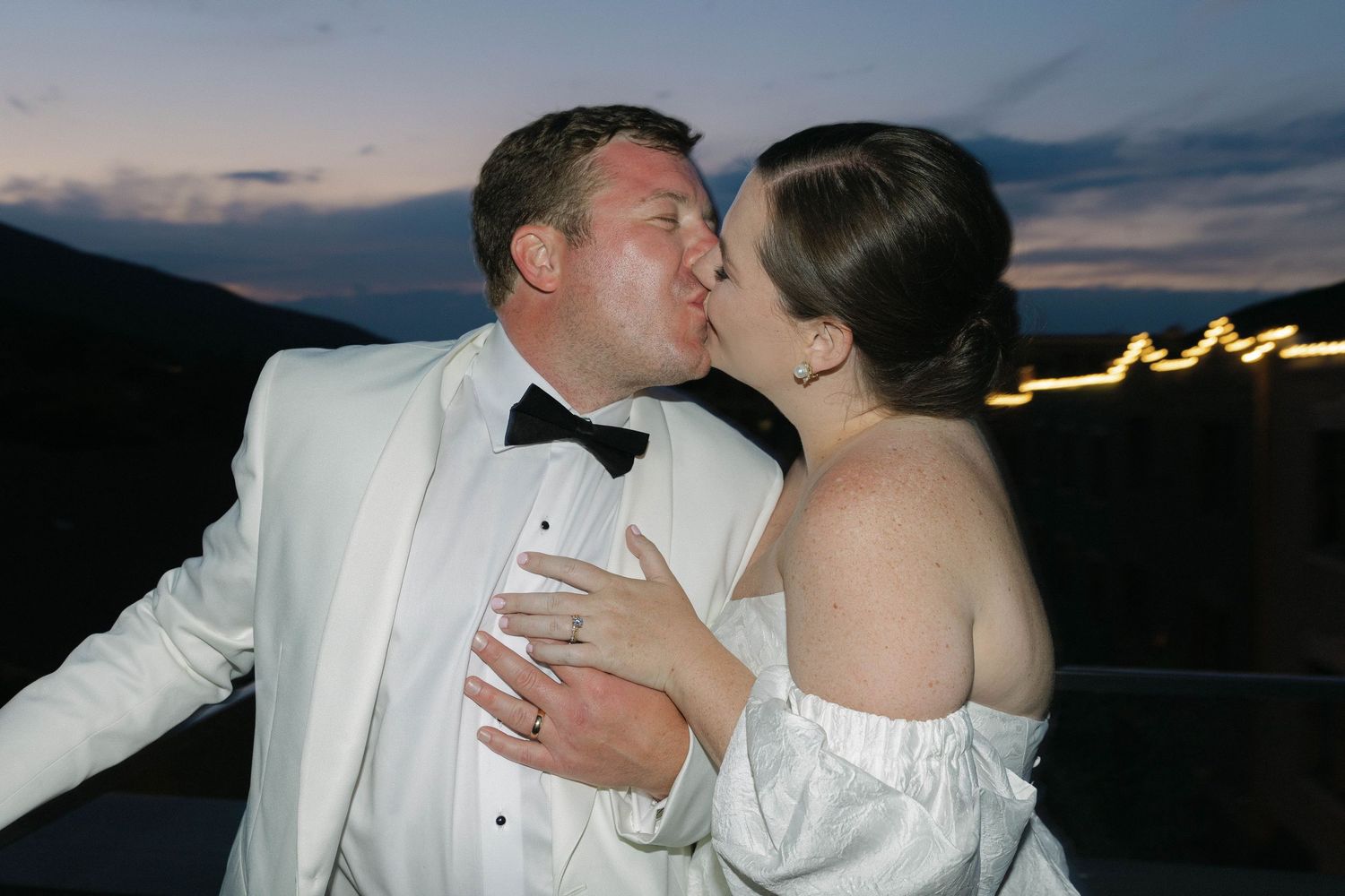 Romantic moment between newlyweds in formal attire against mountain sunset backdrop.