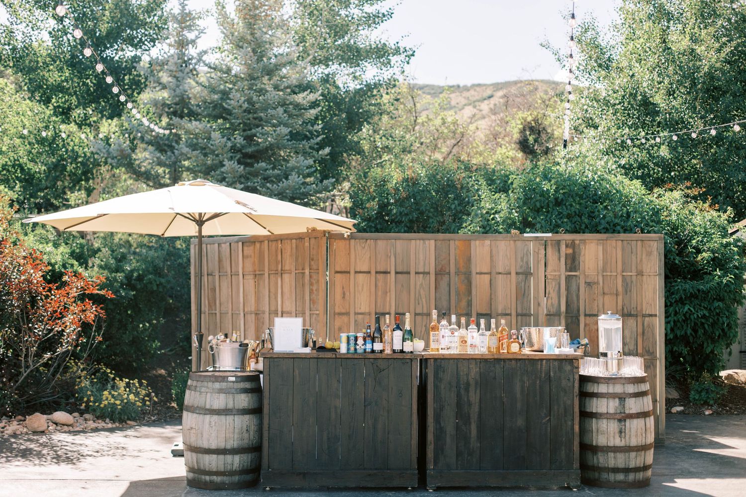 Rustic outdoor bar setup with wooden barrels and umbrella shade at a wedding venue.