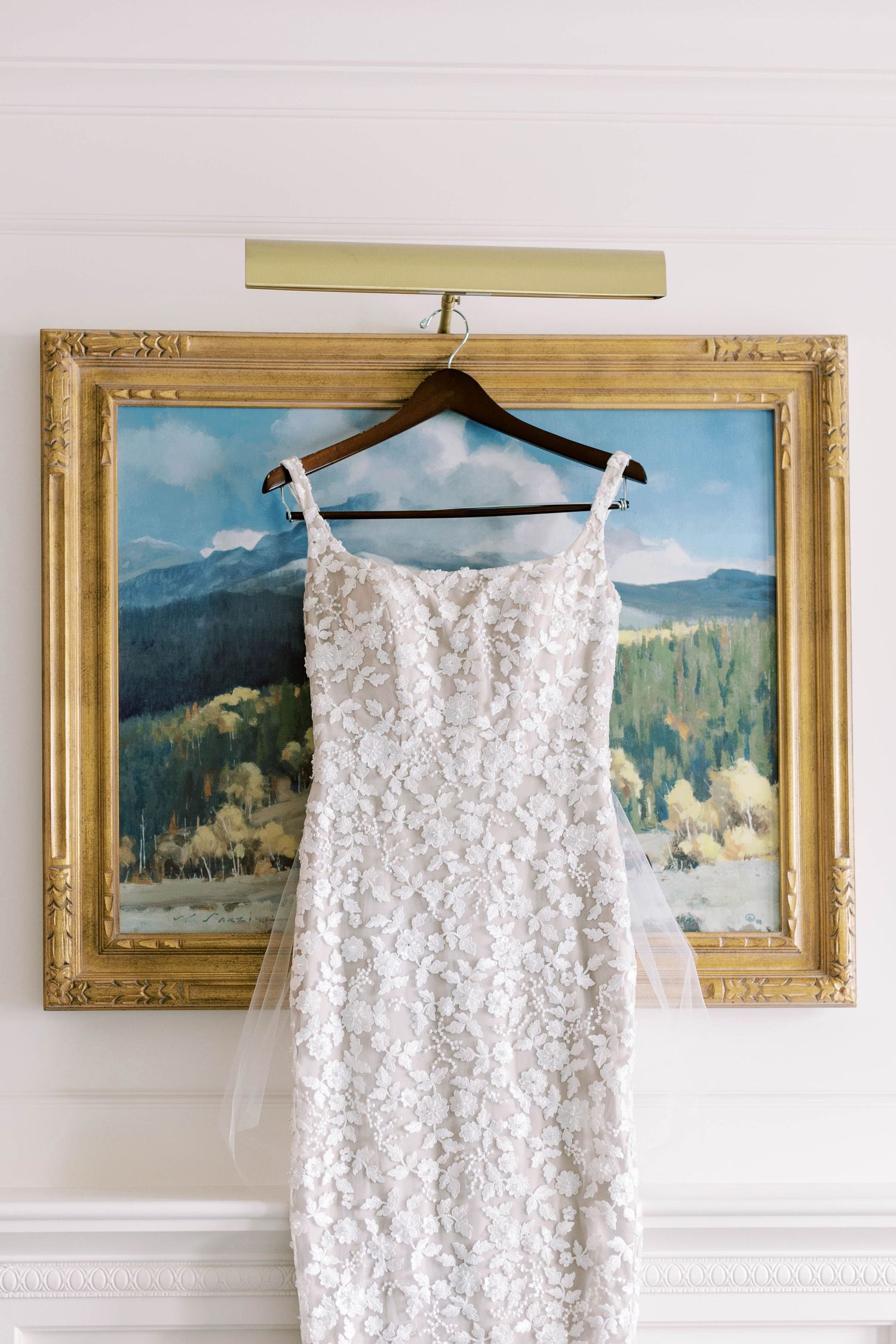 White lace wedding dress hanging on wooden hanger against landscape painting in gold frame.