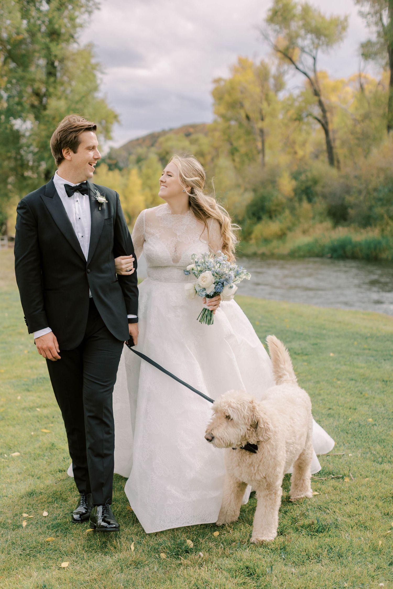 Couple walks their goldendoodle dog wearing bowtie during outdoor wedding photos.