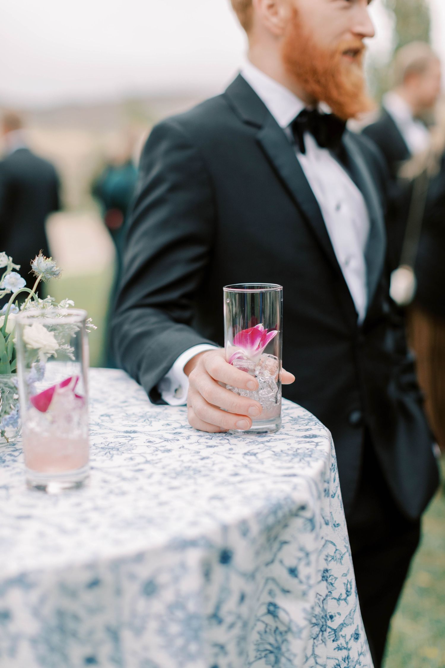 A person in a tuxedo holding a pink cocktail on a lace-covered table at an elegant event.
