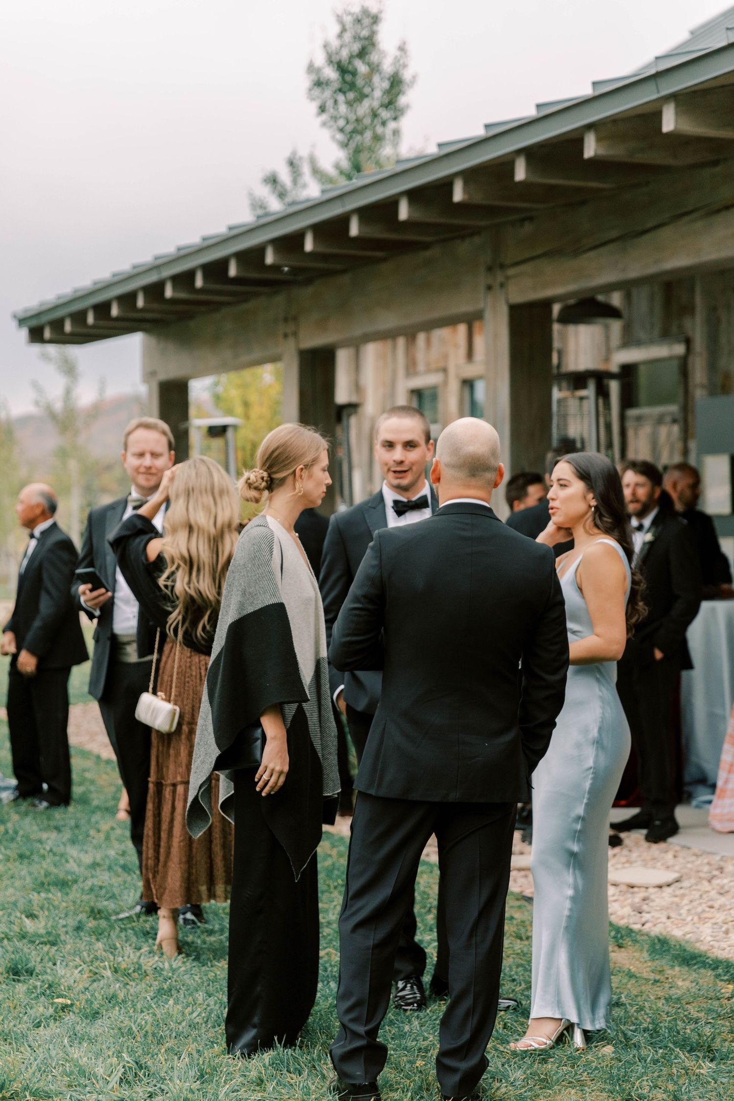 Wedding guests in formal attire gathered outside rustic venue during cocktail hour.