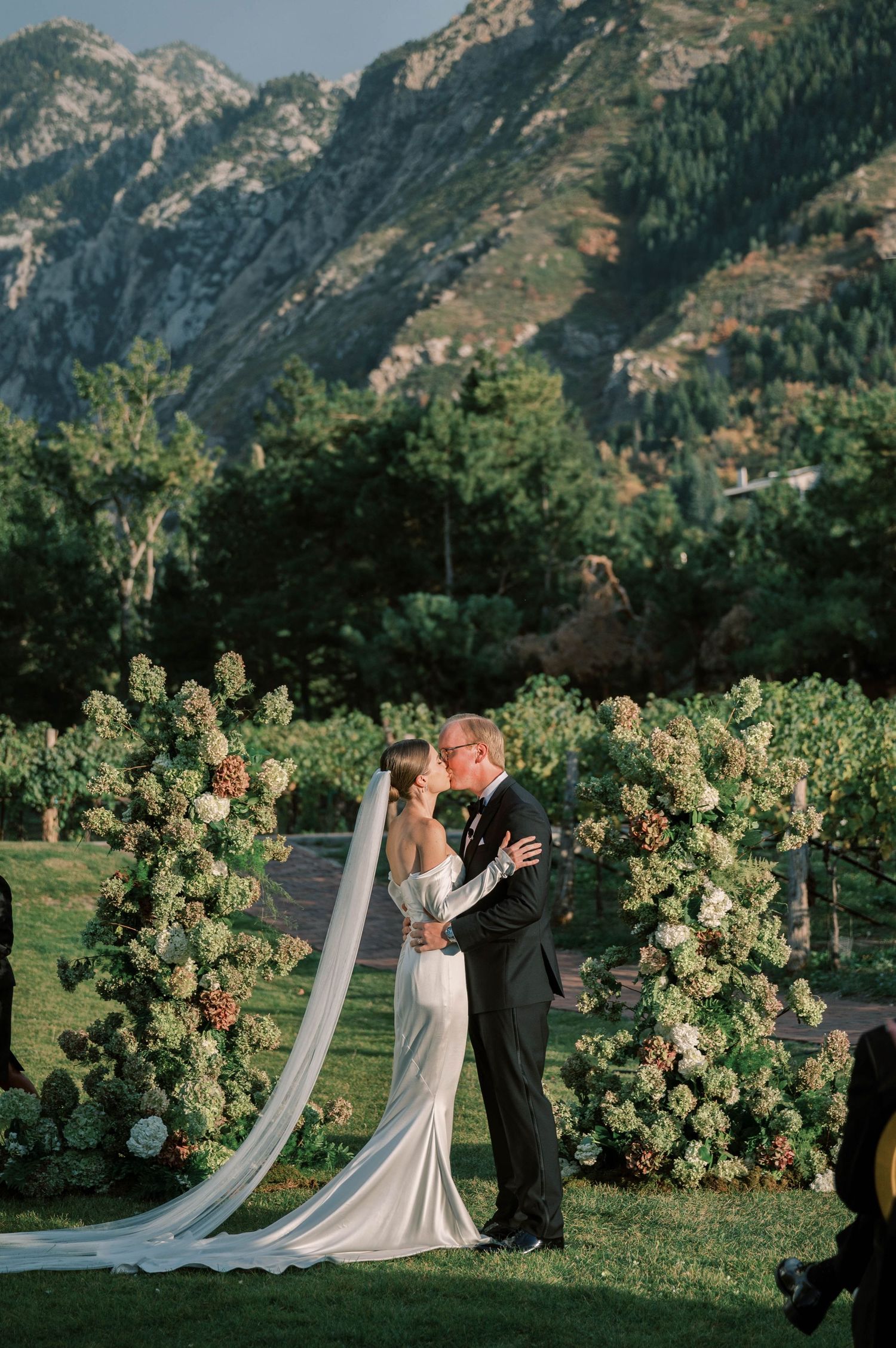 Newlyweds share romantic moment under floral arch with dramatic mountain landscape in background.