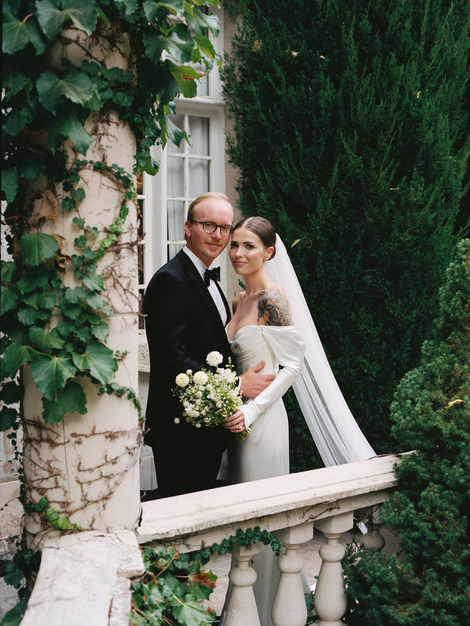 Newlyweds pose with bouquet on an ivy-covered balcony with ornate stone railings.