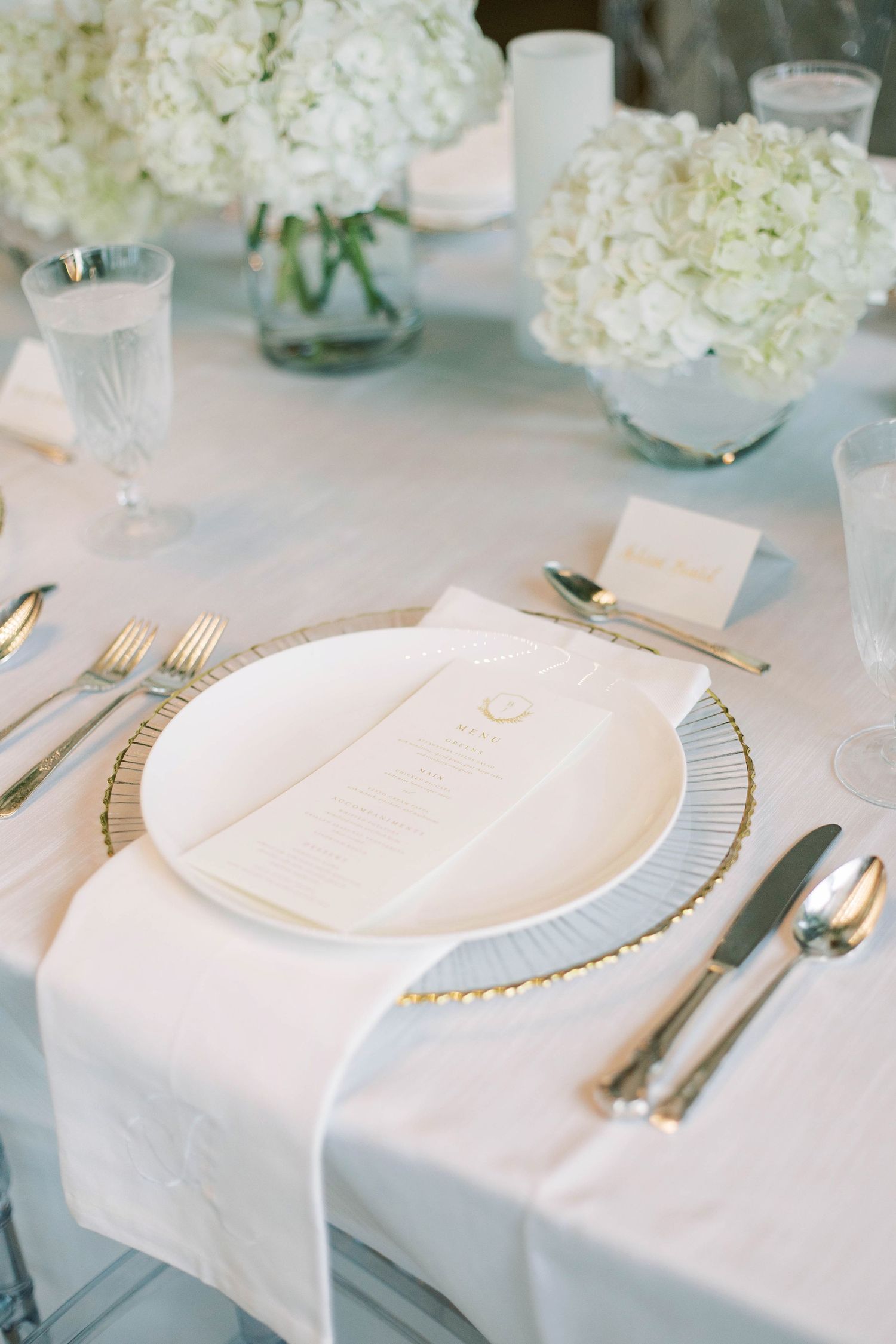 Classic white place setting with gold charger plate and hydrangea centerpiece.