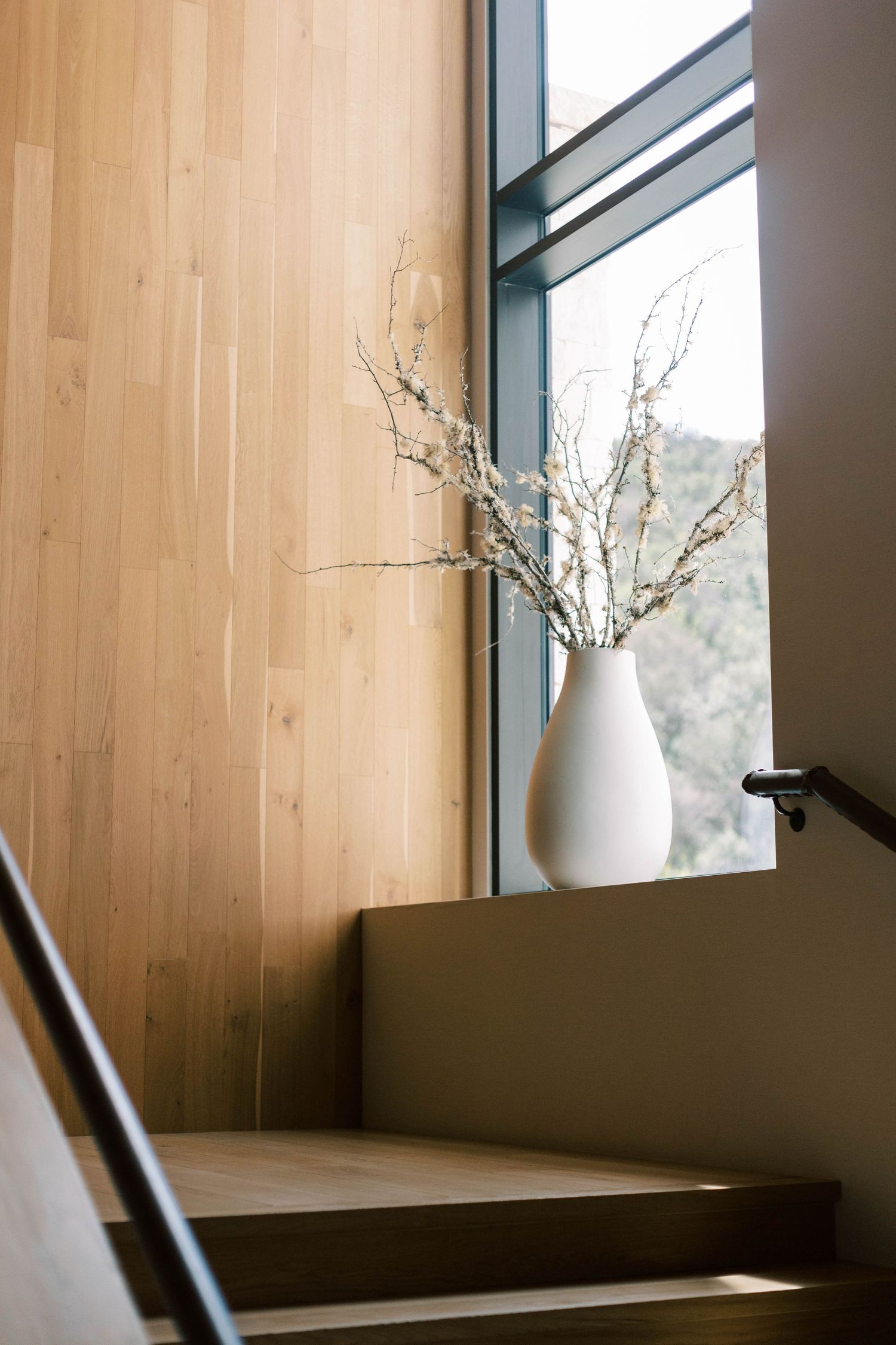 Minimalist vase with dried branches on windowsill with natural wood paneled walls.