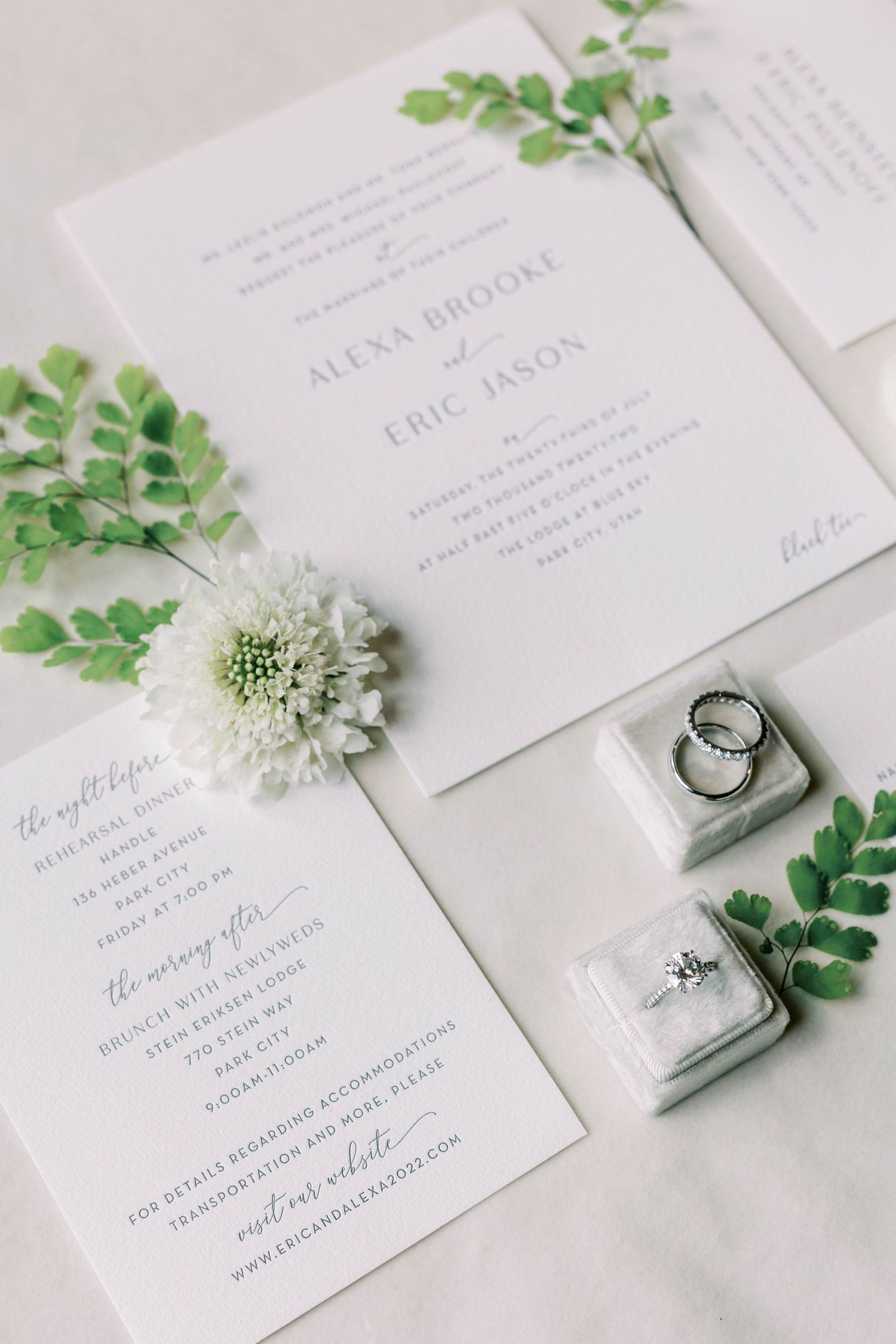 Wedding invitation suite with white floral details, delicate greenery, and wedding rings on a marble ring box.