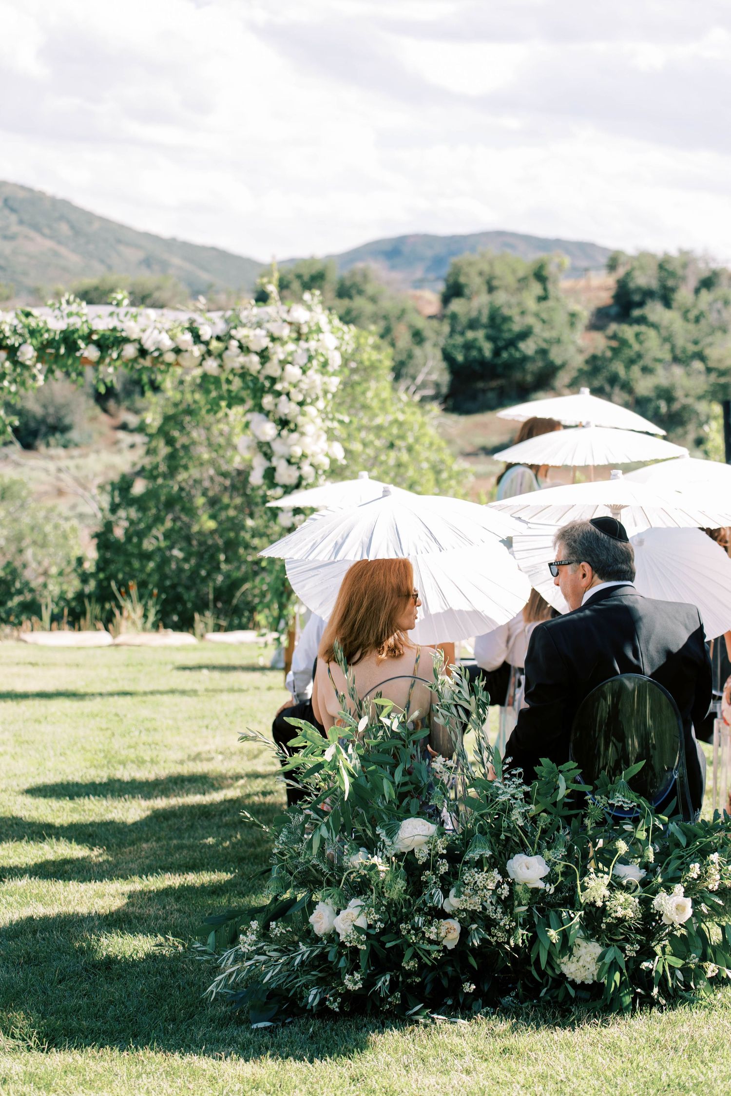 Wedding ceremony seating with white parasols and floral arrangements in a mountain setting.
