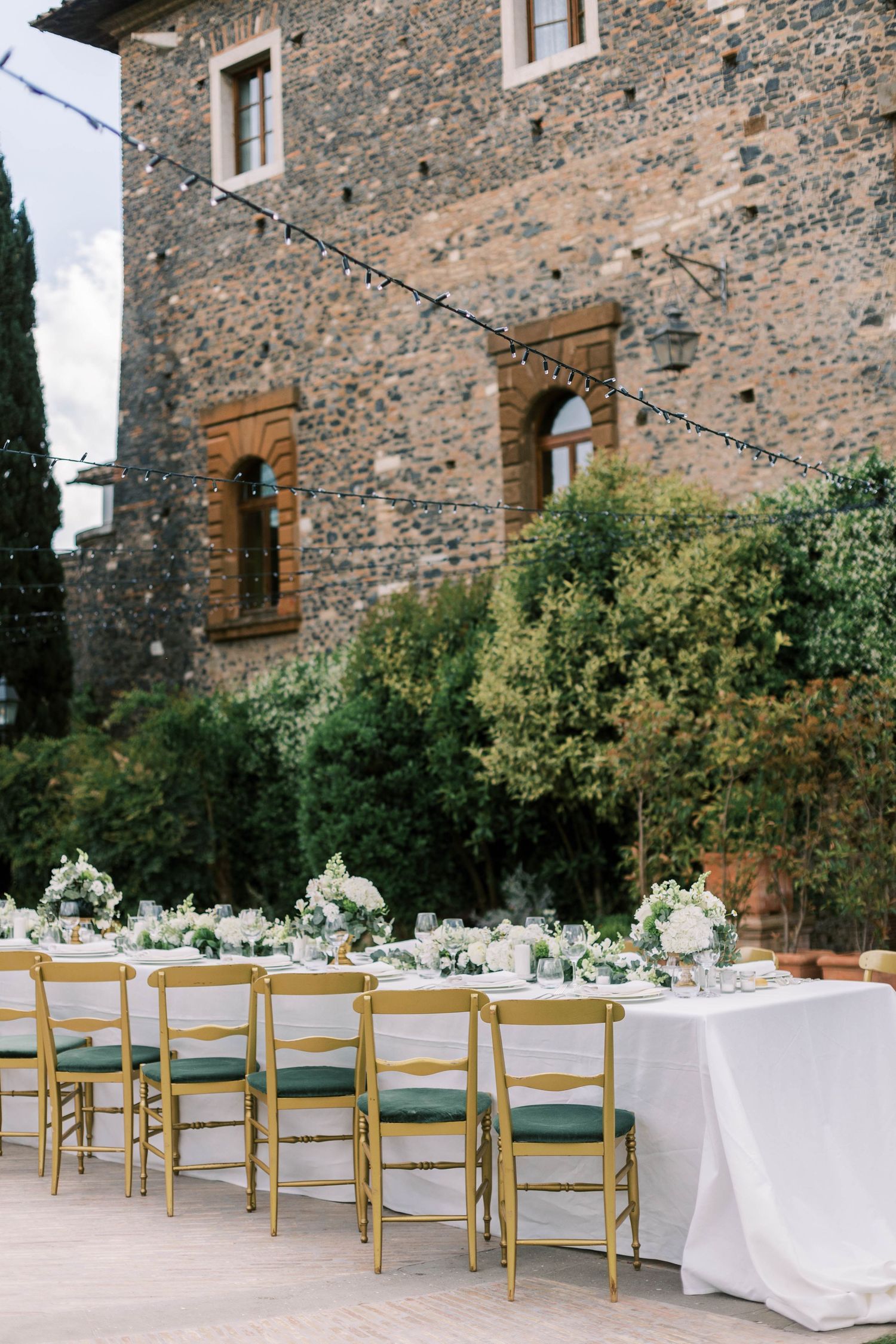 Long wedding reception table with white linens and gold chairs against stone villa facade at castello della castellucia in rome italy.