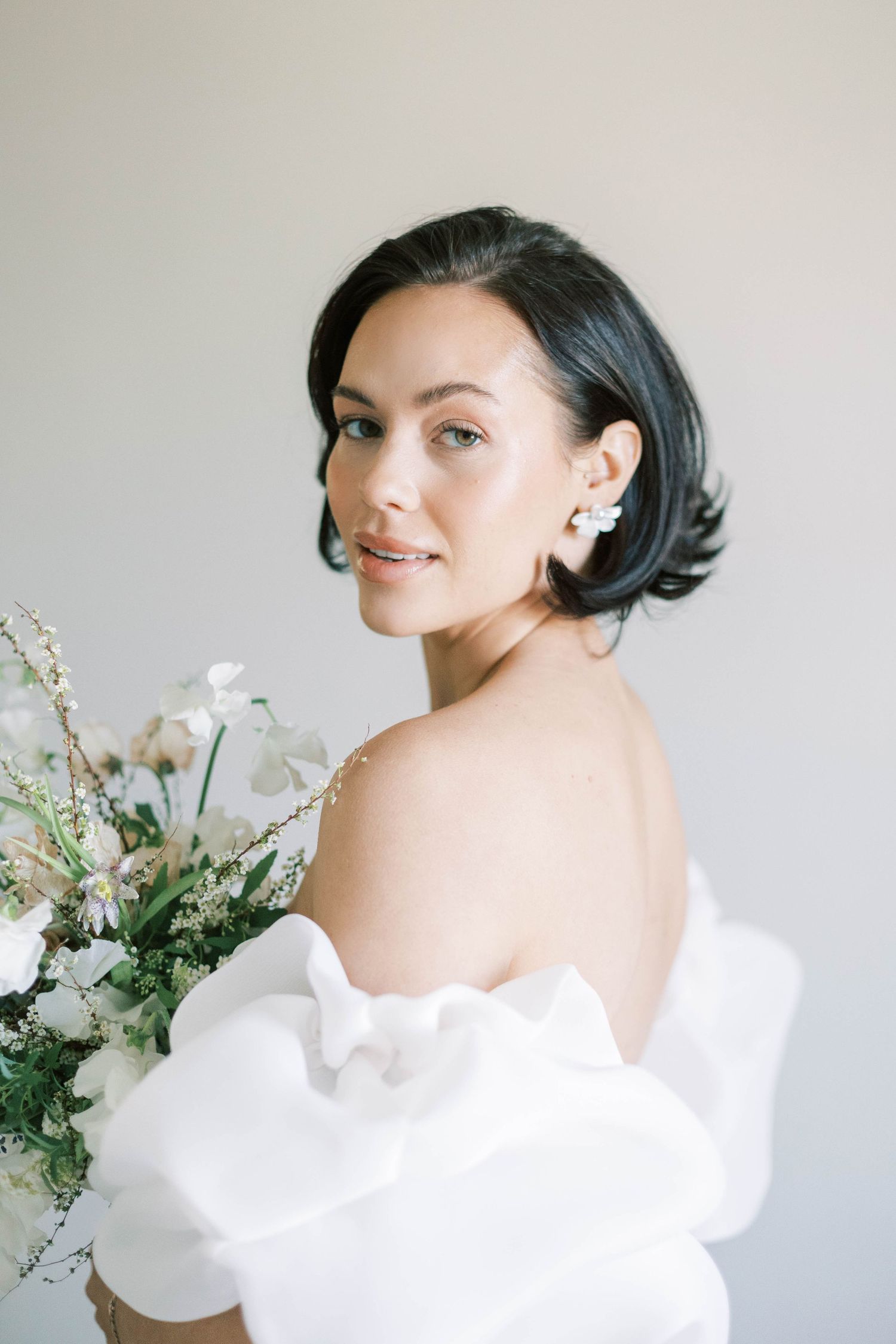 Elegant bride wearing an off-shoulder white dress holds a delicate bouquet of white flowers and greenery against neutral background.