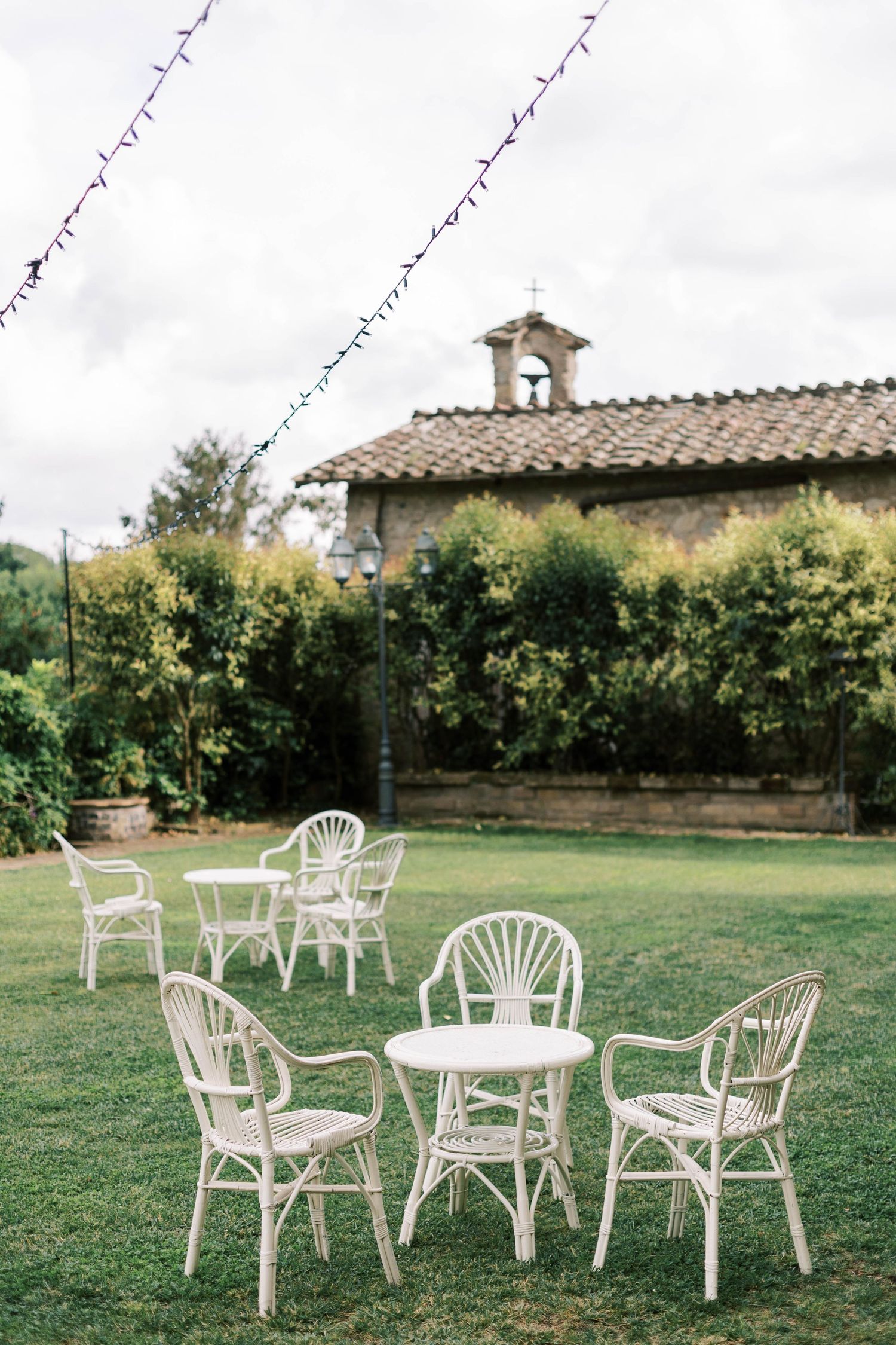 White garden furniture arranged on lawn with string lights and chapel in background.