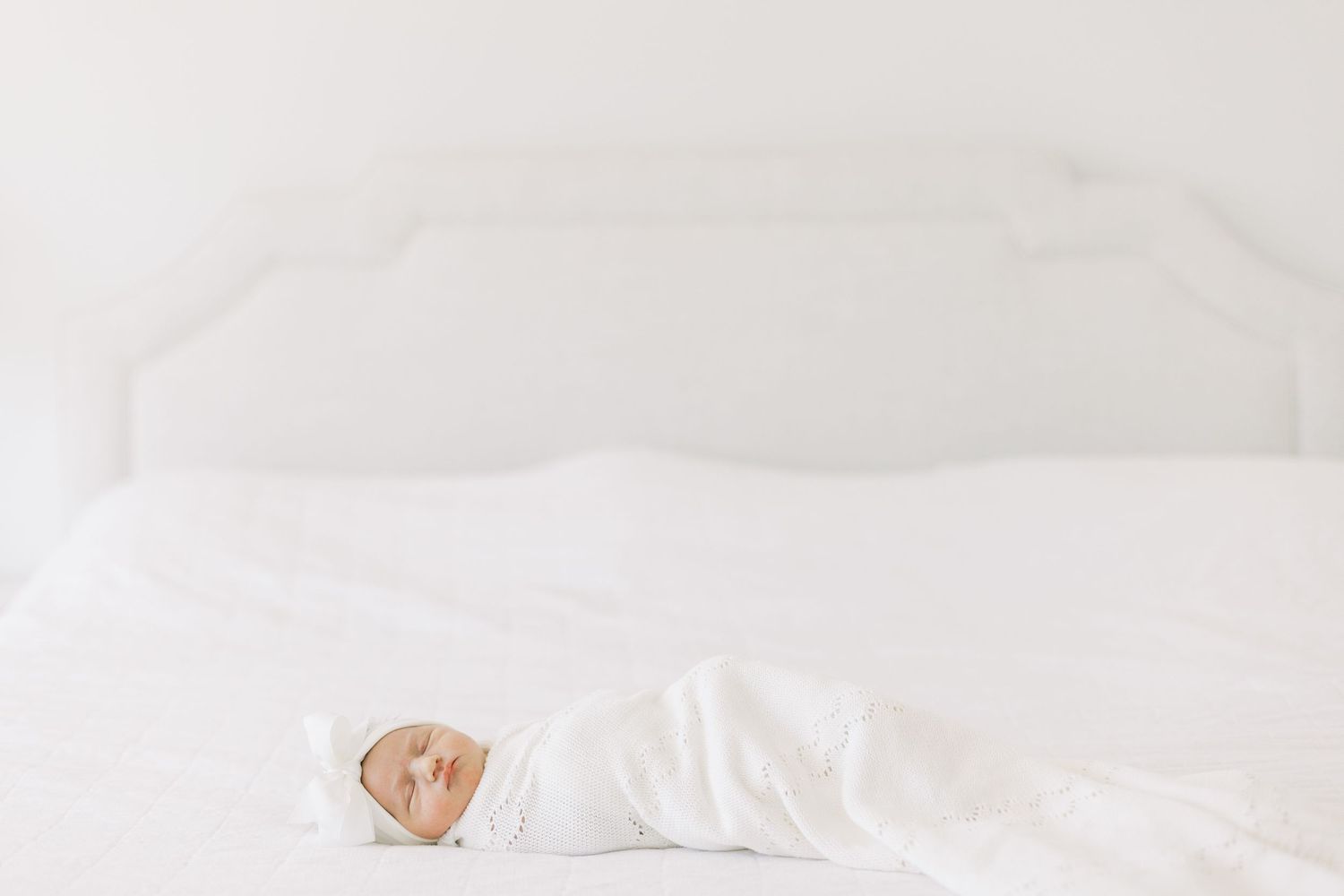 Newborn baby wrapped in white swaddle lying peacefully on white bedding.