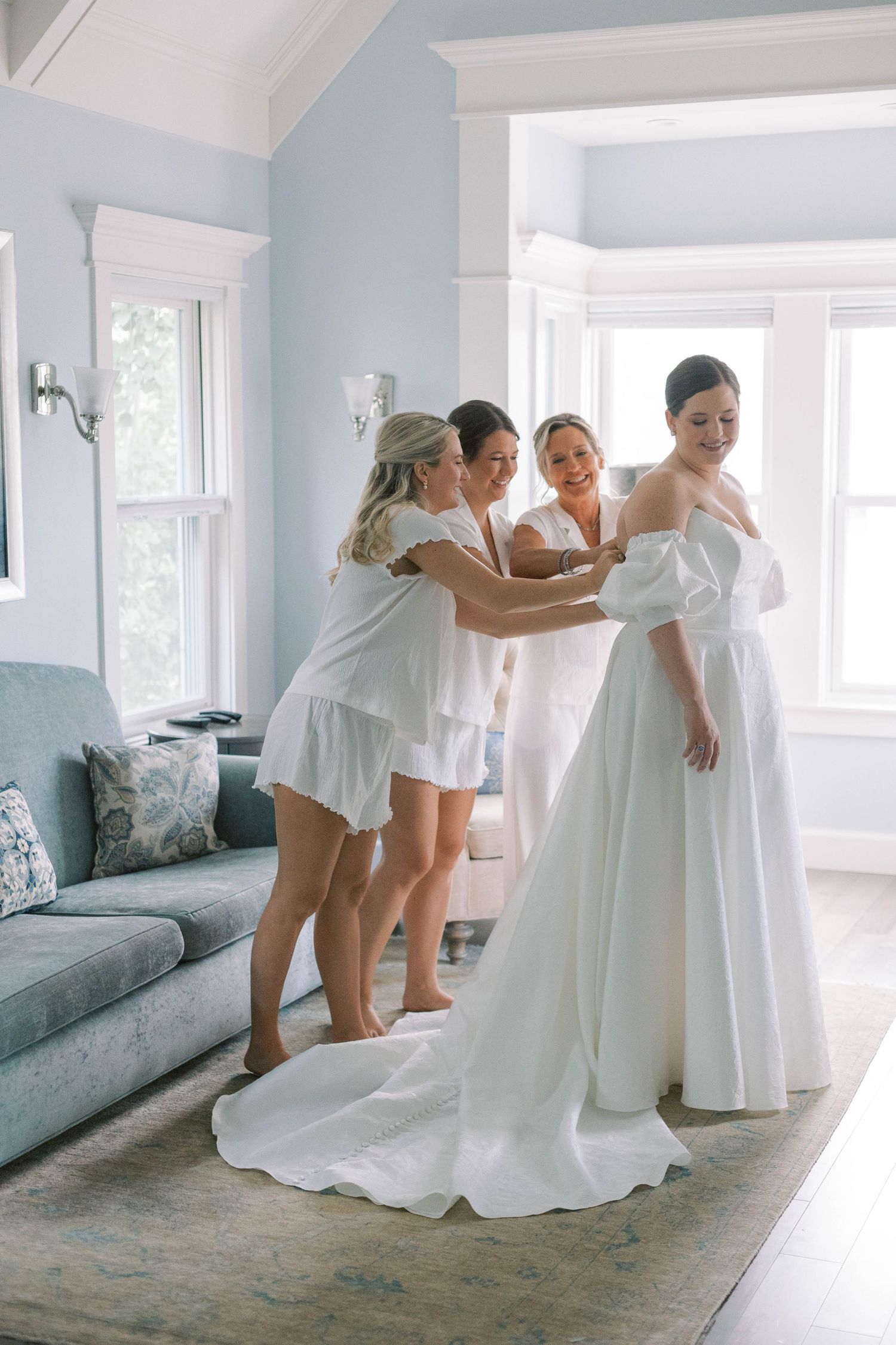 Bridesmaids in white dresses help the bride get ready in a bright room with blue walls.