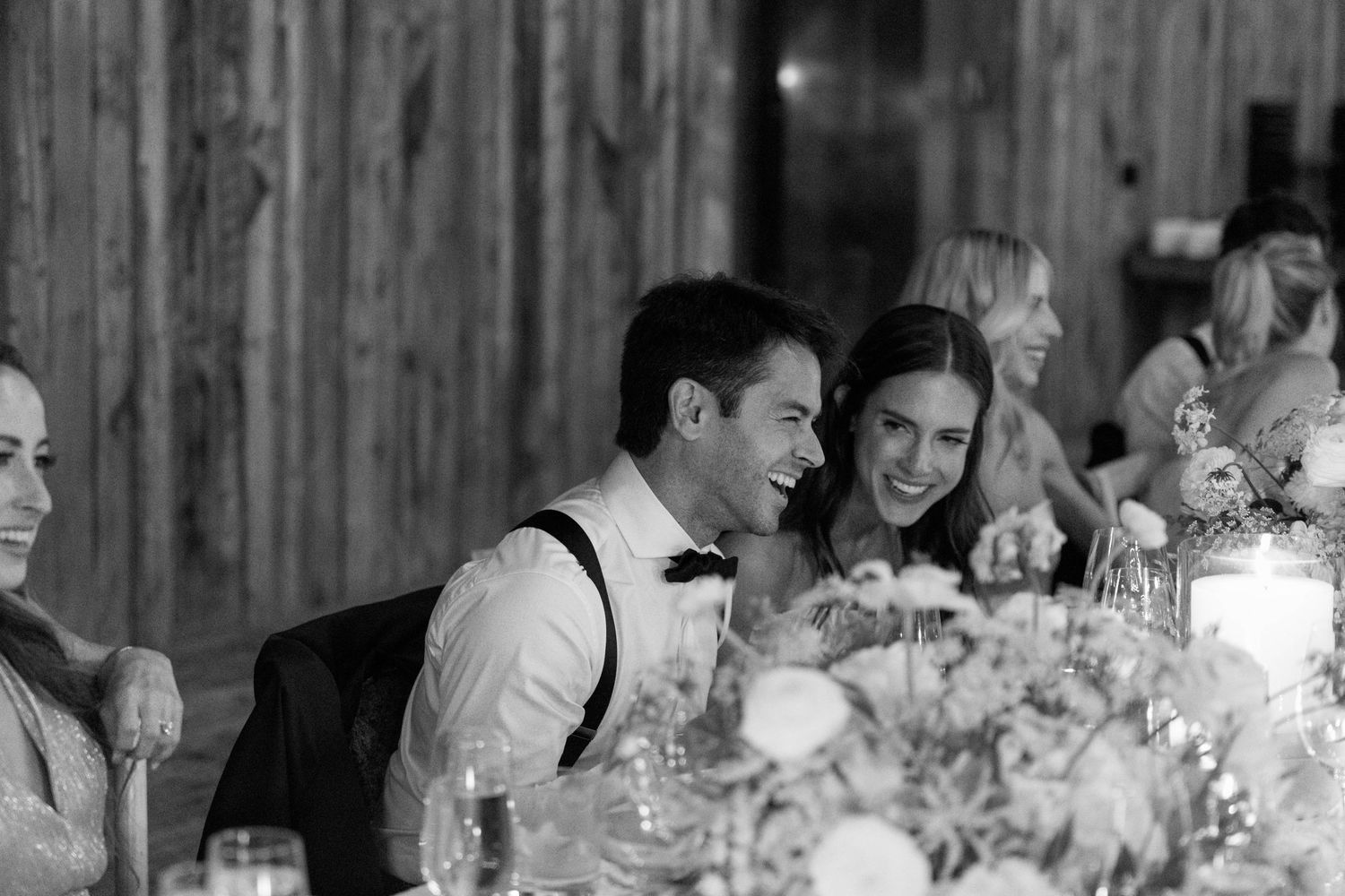 Candlelit wedding reception with guests laughing at head table in rustic barn.