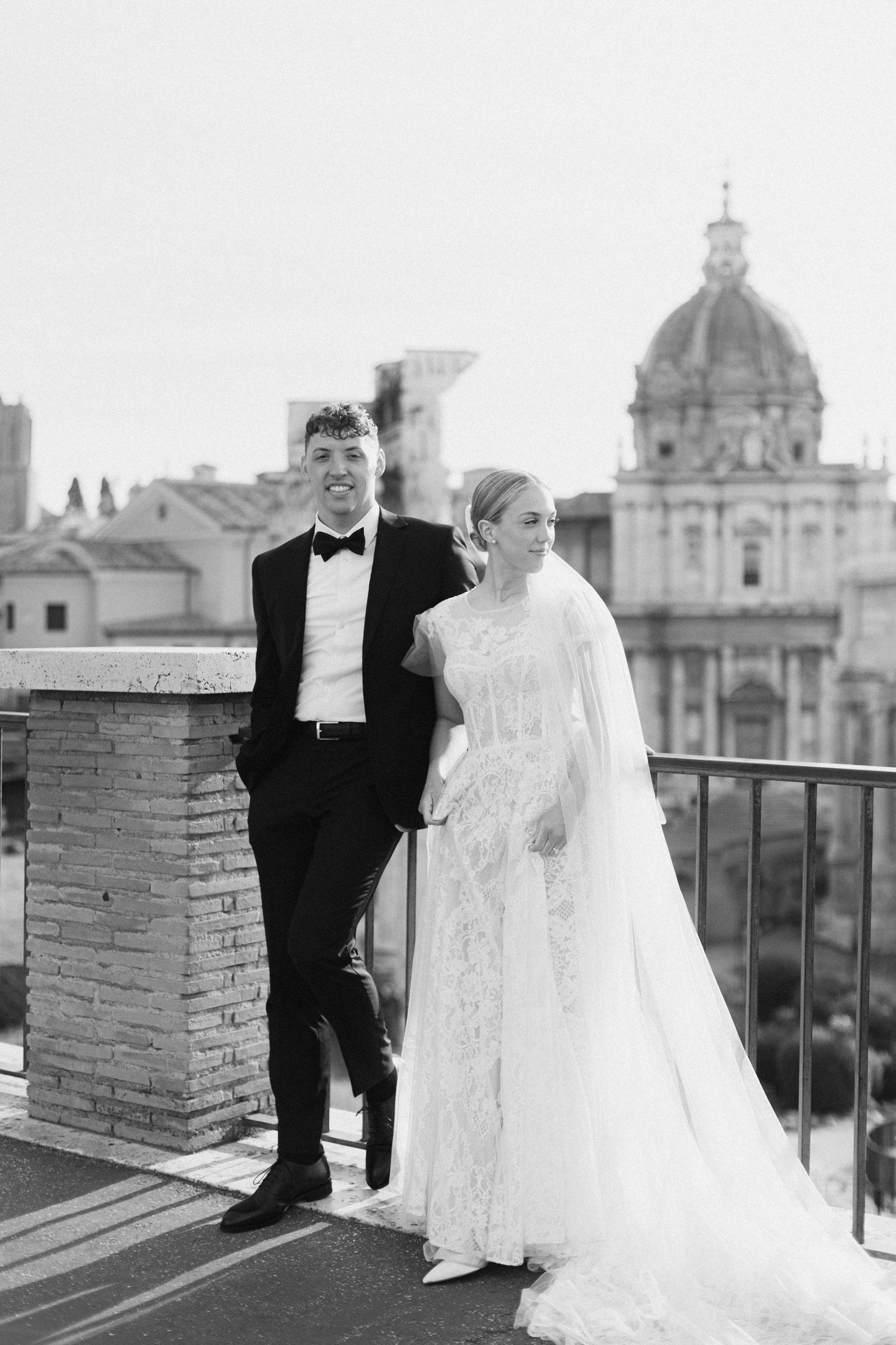 Elegant couple poses on rooftop terrace with Rome's historic dome architecture in background.