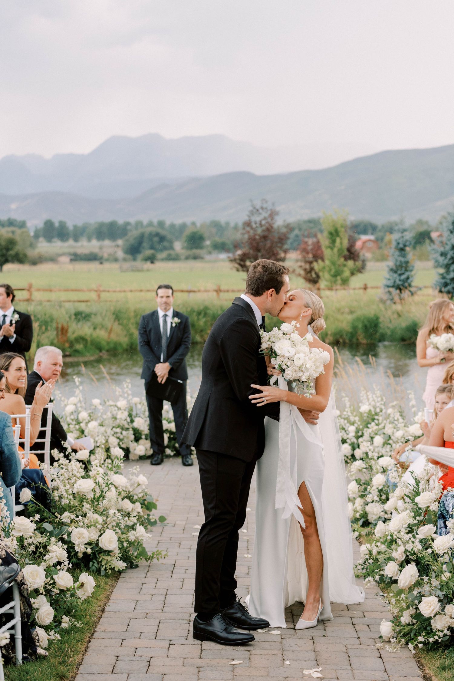 Newlyweds share first kiss on stone pathway lined with white floral arrangements and mountain scenery.