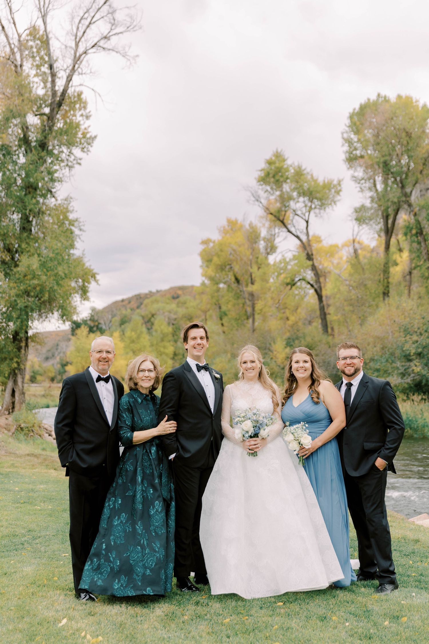 Small wedding party group poses together outdoors near river with fall foliage in background.