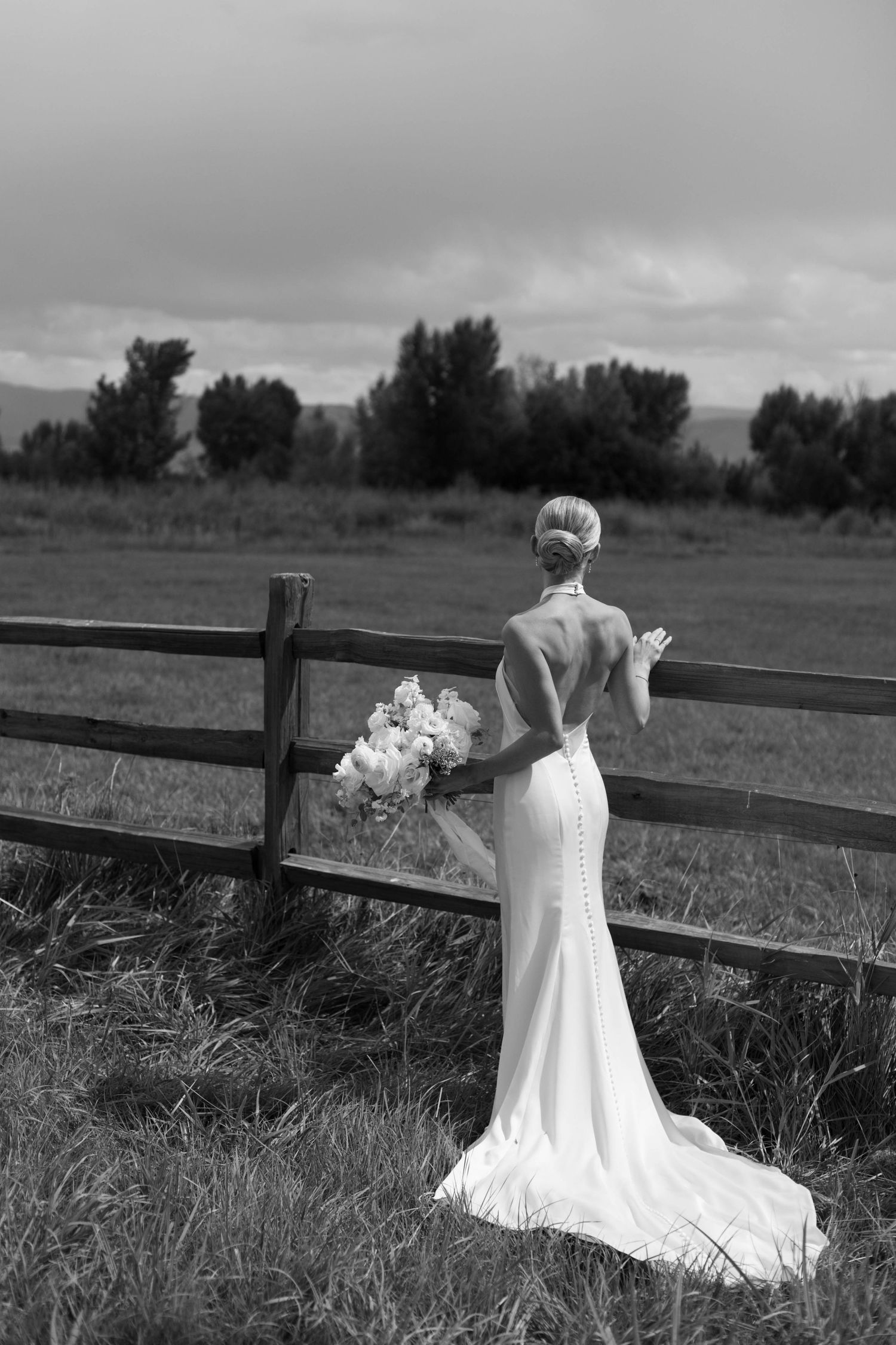 Black and white photo of bride at rustic fence with mountain view.