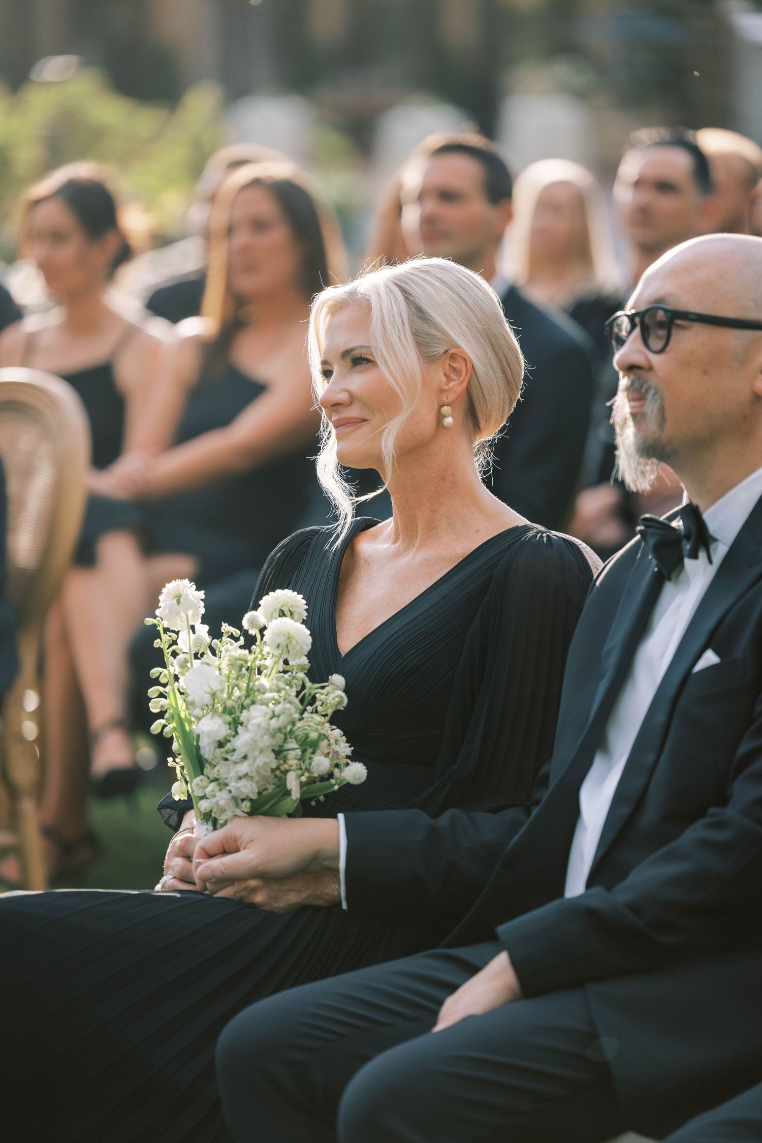 Wedding guests in formal black attire watching ceremony while holding white floral bouquet.