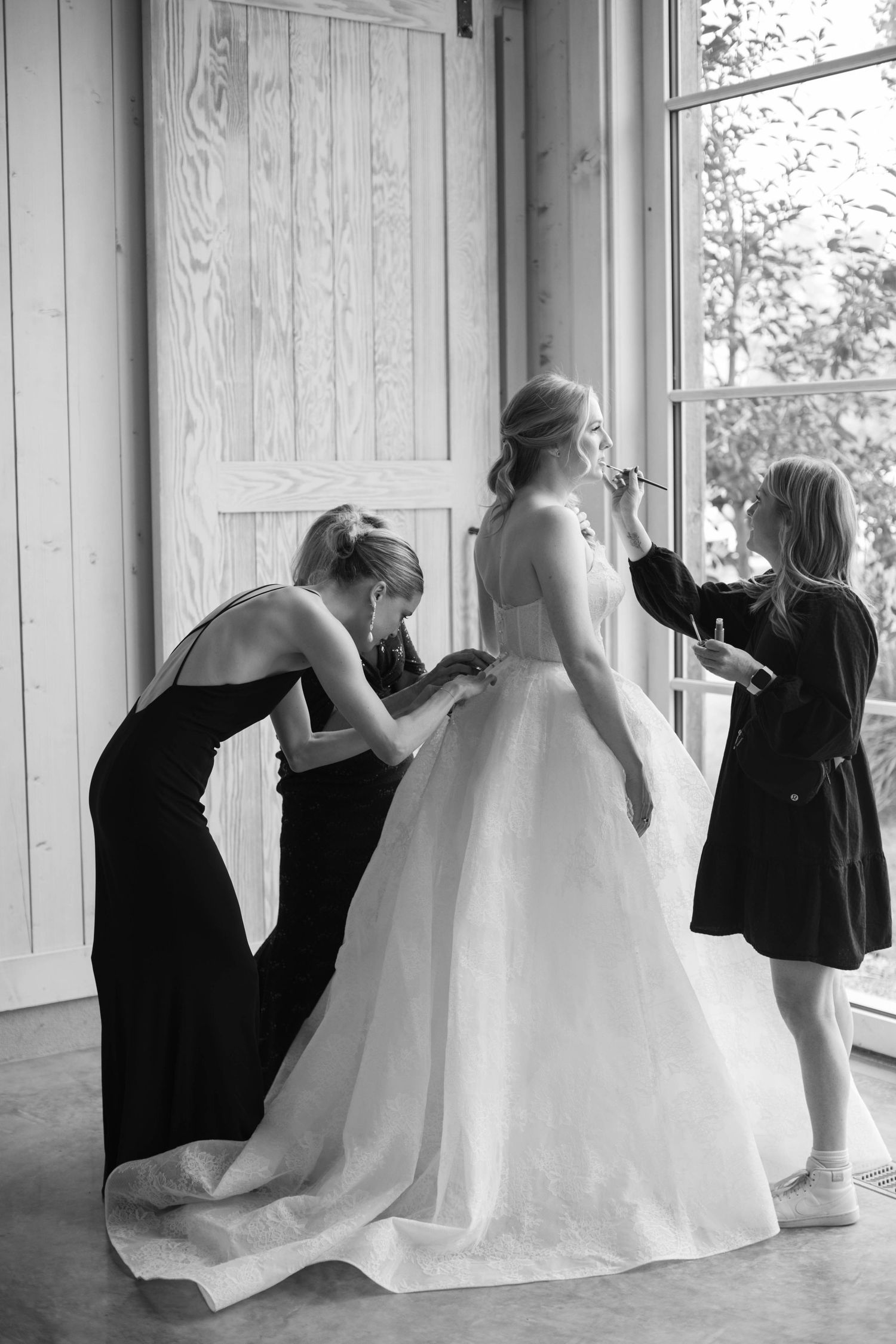 Black and white photo of bridesmaids helping bride with wedding dress near a window.