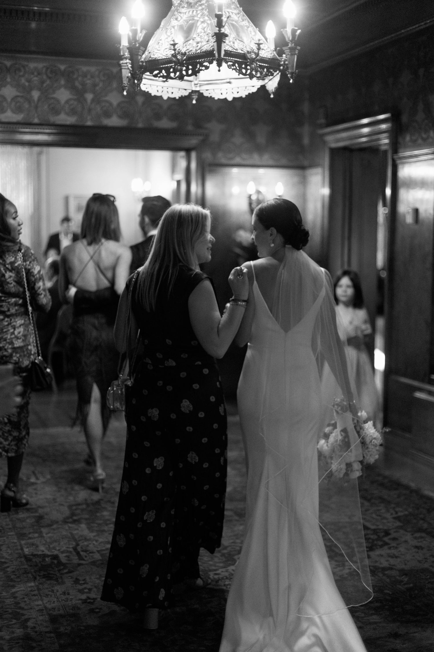 Black and white photo of guests mingling in elegant hallway with chandelier overhead.