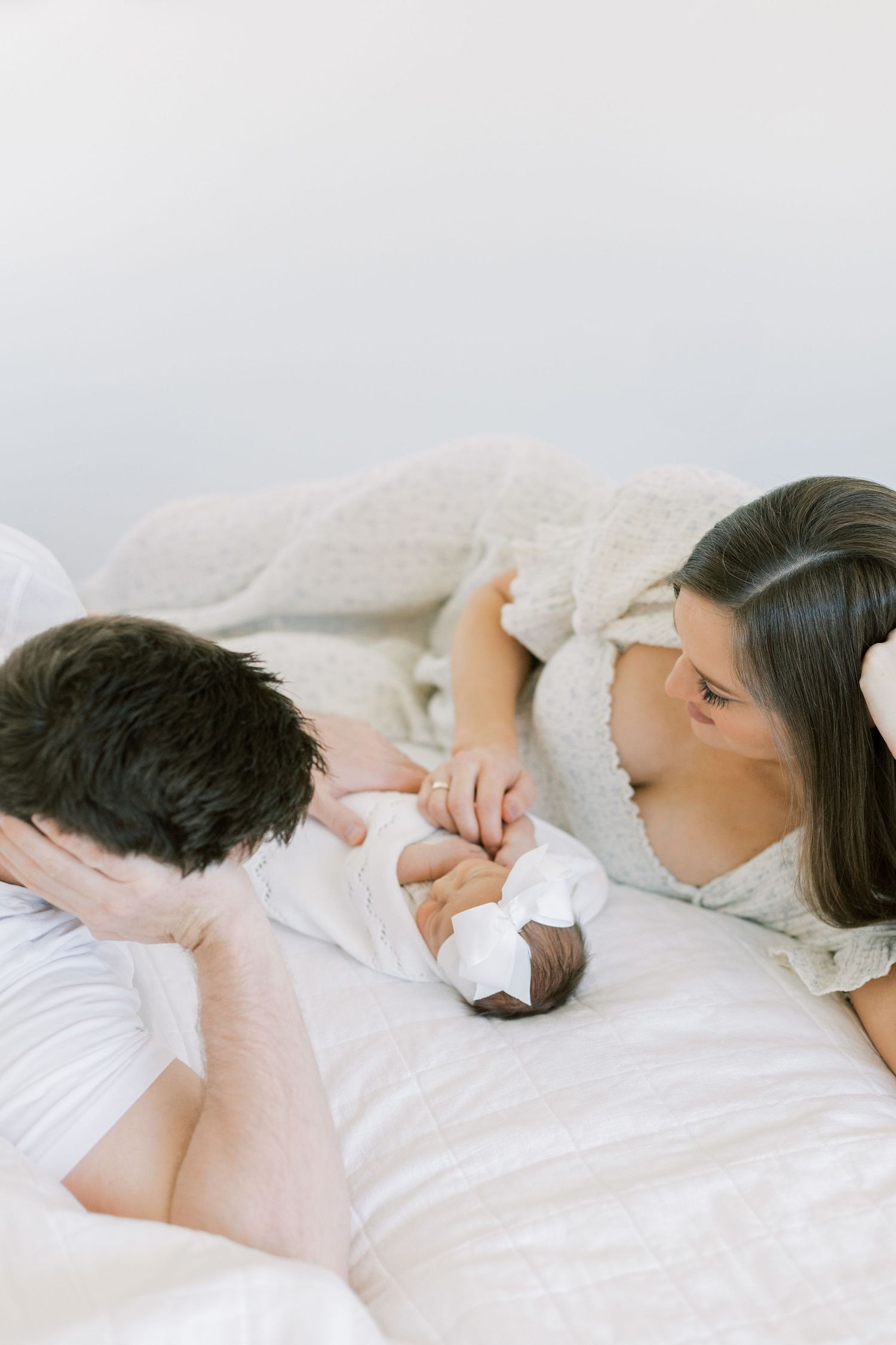 Family lying on white bedding admiring their newborn baby together.