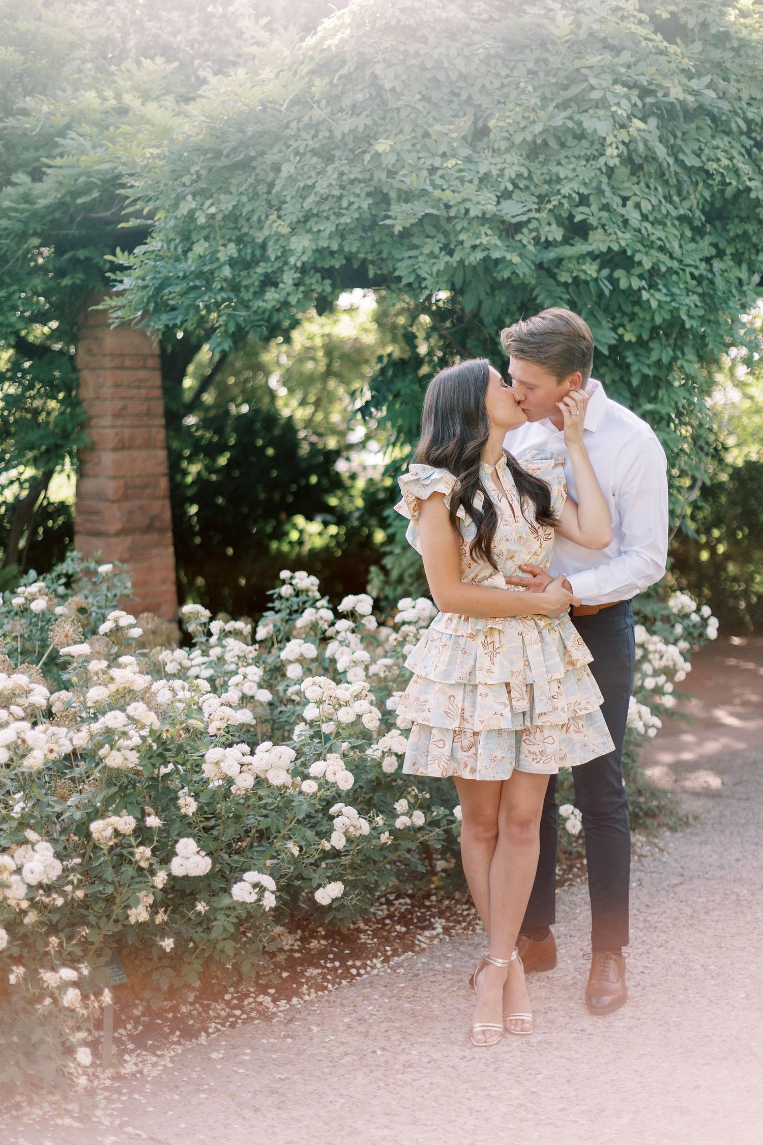 A couple shares an intimate moment among white roses in a garden pathway.