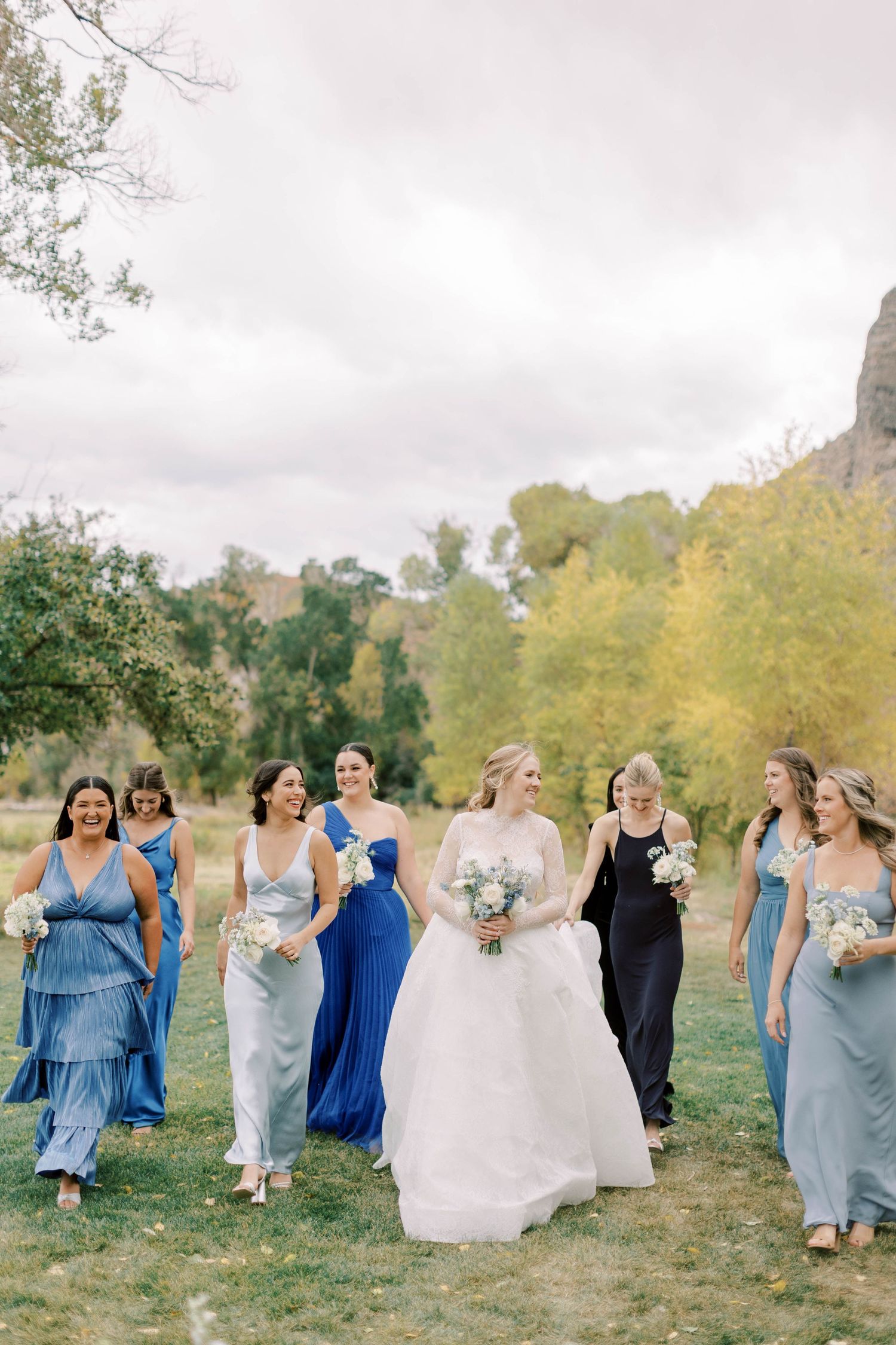 Wedding party in mix of blue and neutral dresses walk together on grassy path with trees in background.