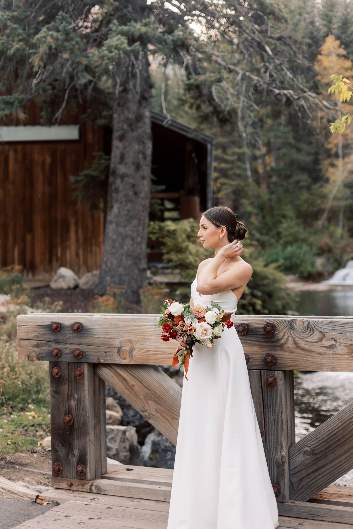 Bride in white dress holding bouquet while standing on wooden bridge in forest setting.