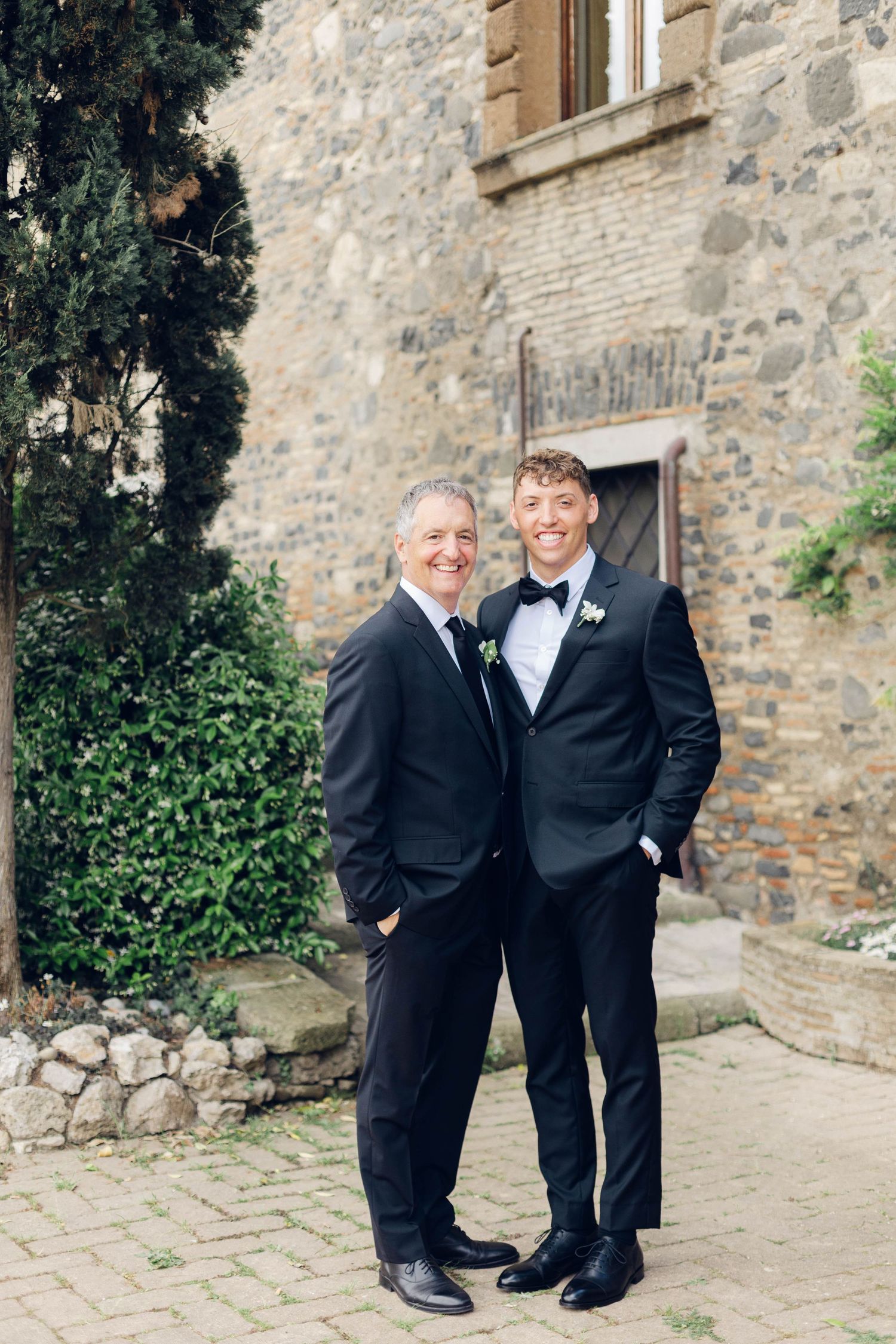 Two men in dark suits share joyful moment outside stone building.