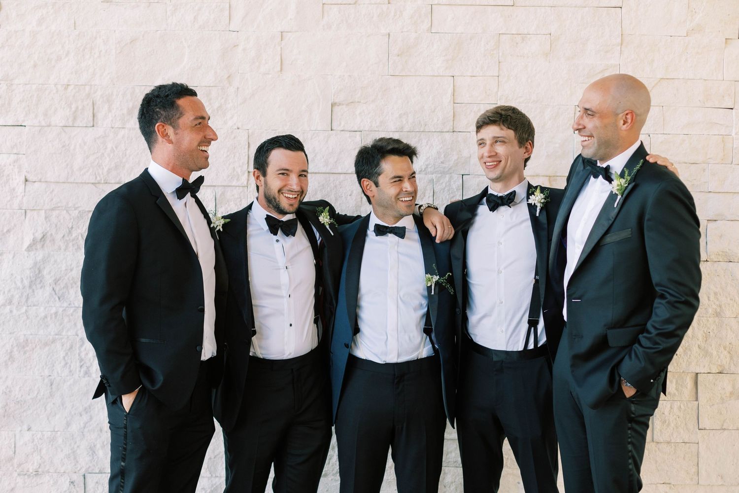 Five groomsmen in black tuxedos and bowties pose casually against white stone wall.
