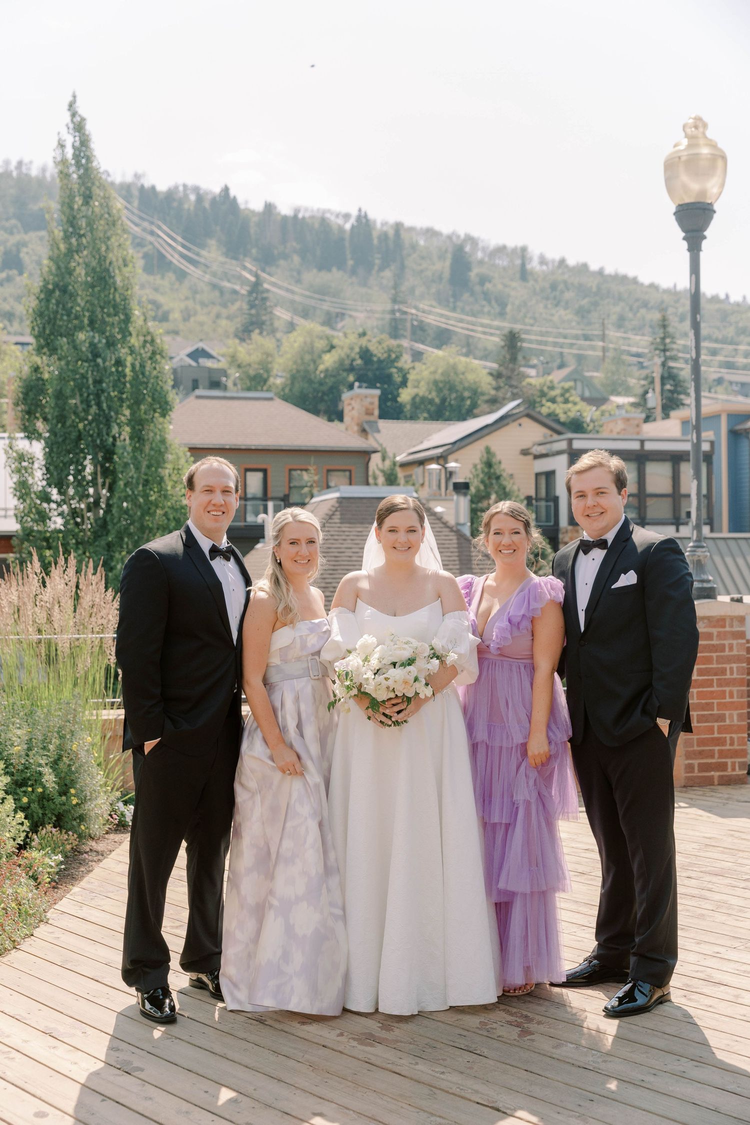 Wedding party poses on wooden deck with mountain town backdrop.