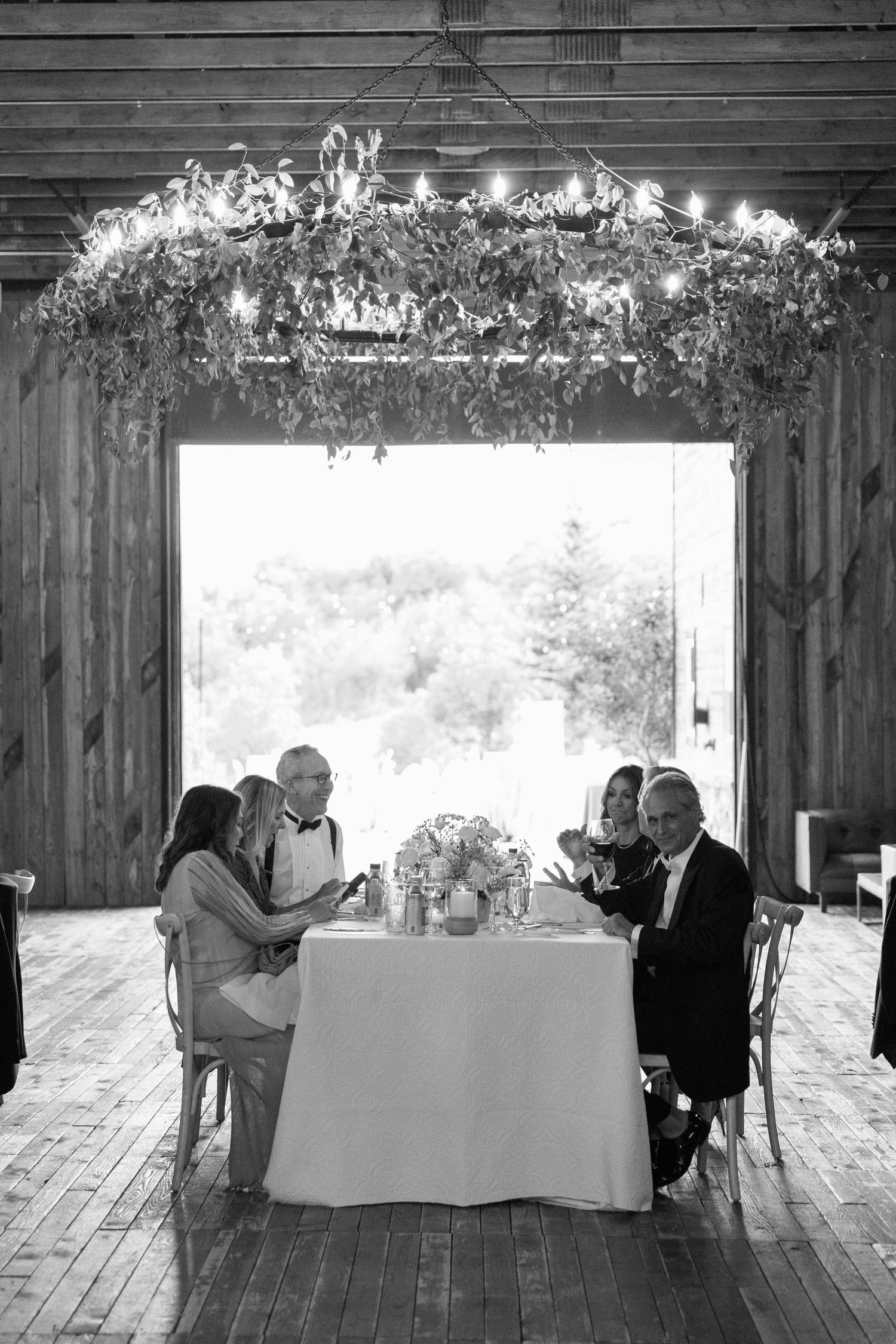 Intimate wedding reception in a rustic barn with string lights and greenery overhead.