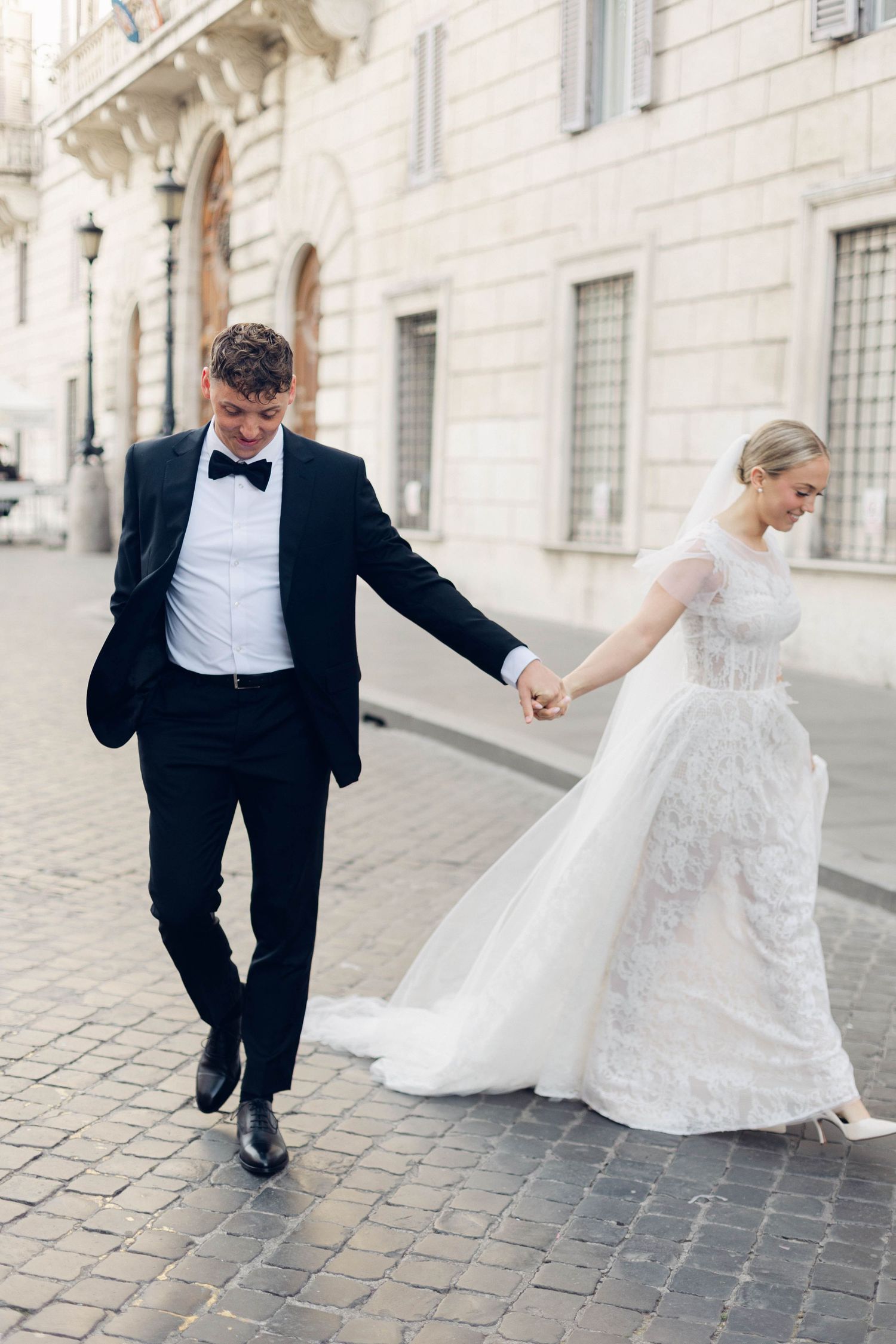 A wedding couple walks playfully together on cobblestone streets with the bride's white gown flowing elegantly behind them.
