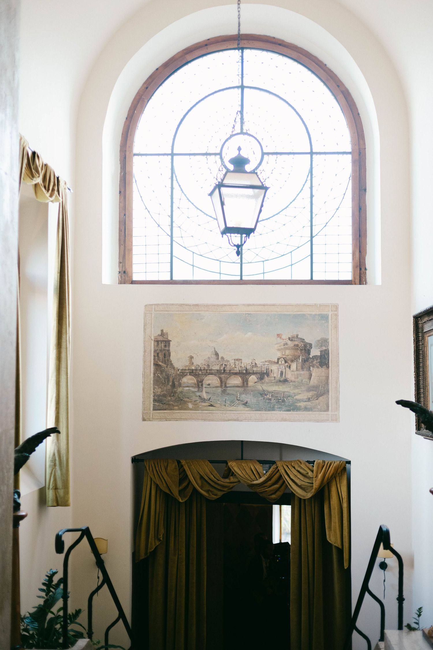 Elegant arched window with lantern and vintage artwork above dark curtains in a historic interior of castello della castellucia in rome.
