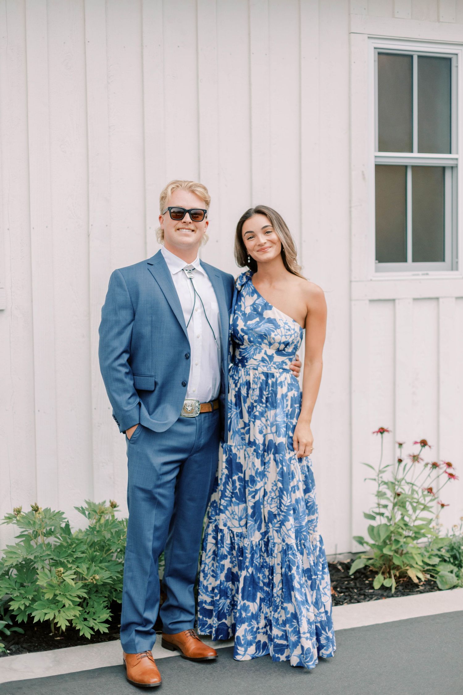 Couple in formal attire posing against white building exterior.