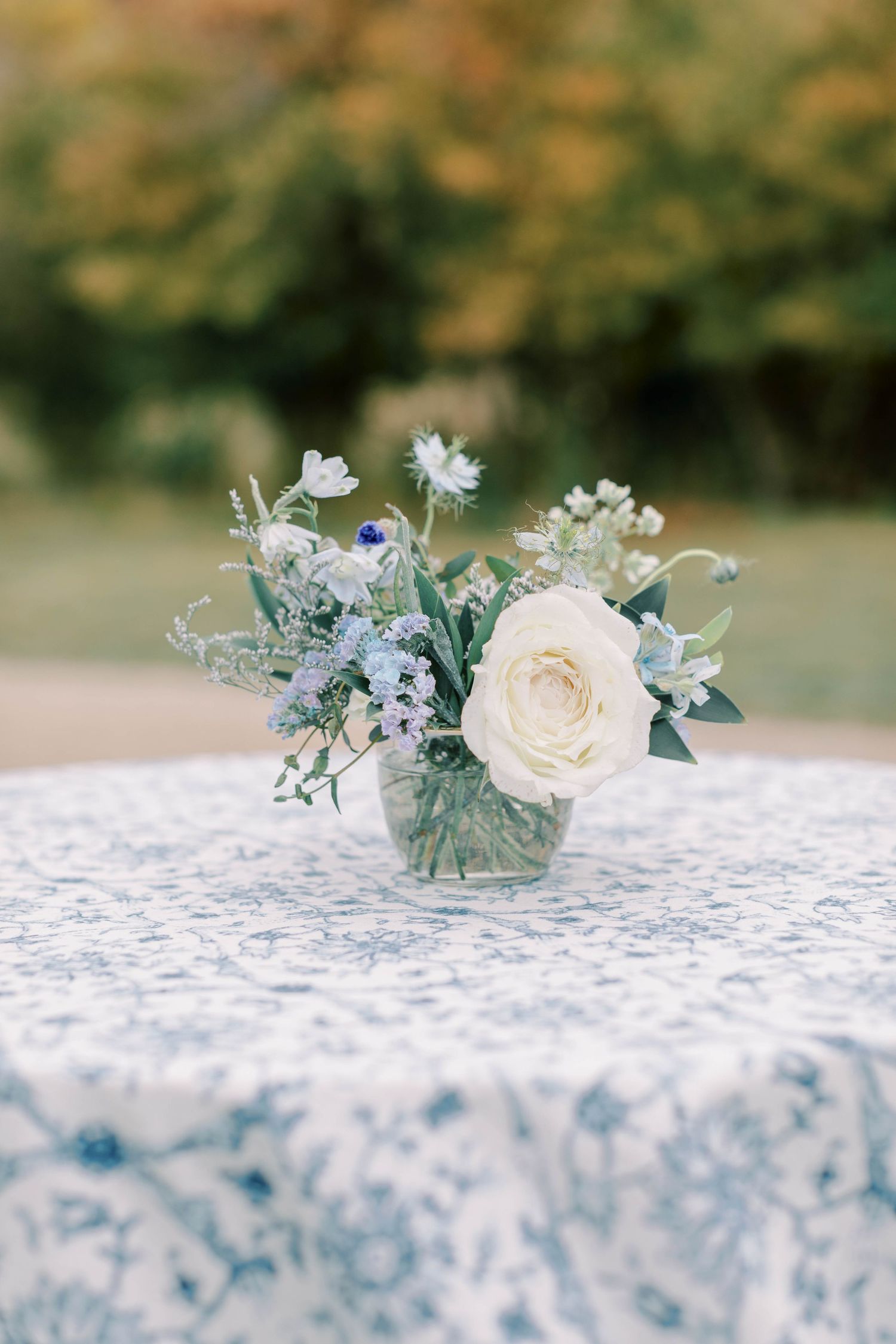 Delicate floral centerpiece with white roses and blue flowers in small glass vase on patterned tablecloth.