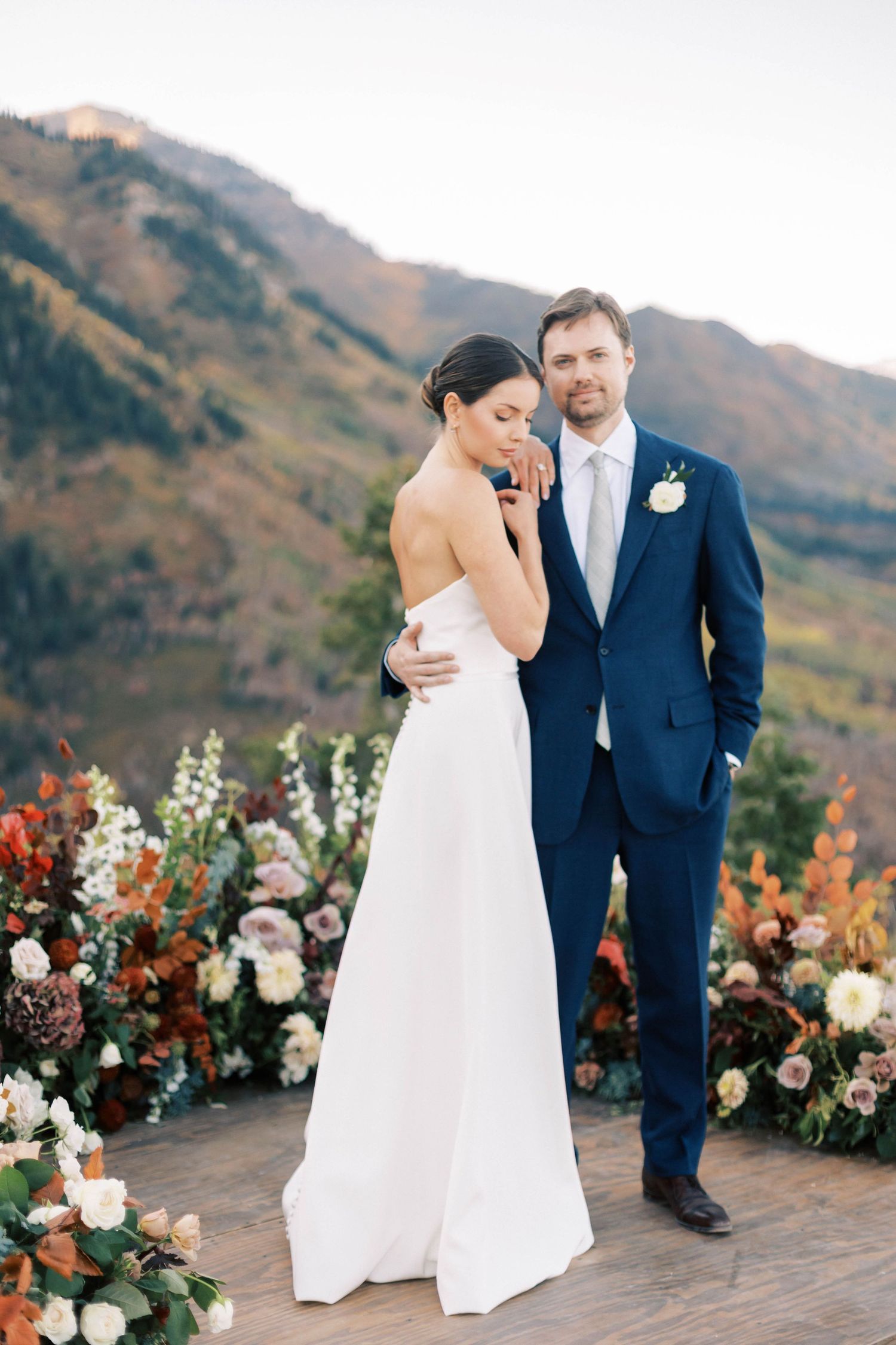 Elegant wedding scene with couple standing by floral arrangements in front of mountain scenery.