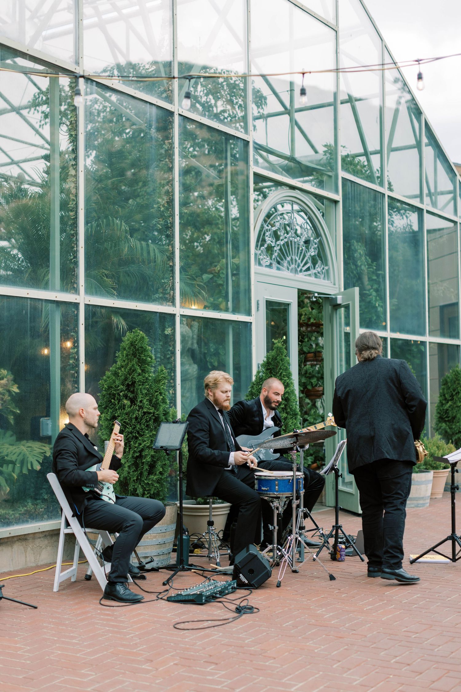 Live band performing outside glass greenhouse venue with string lights.